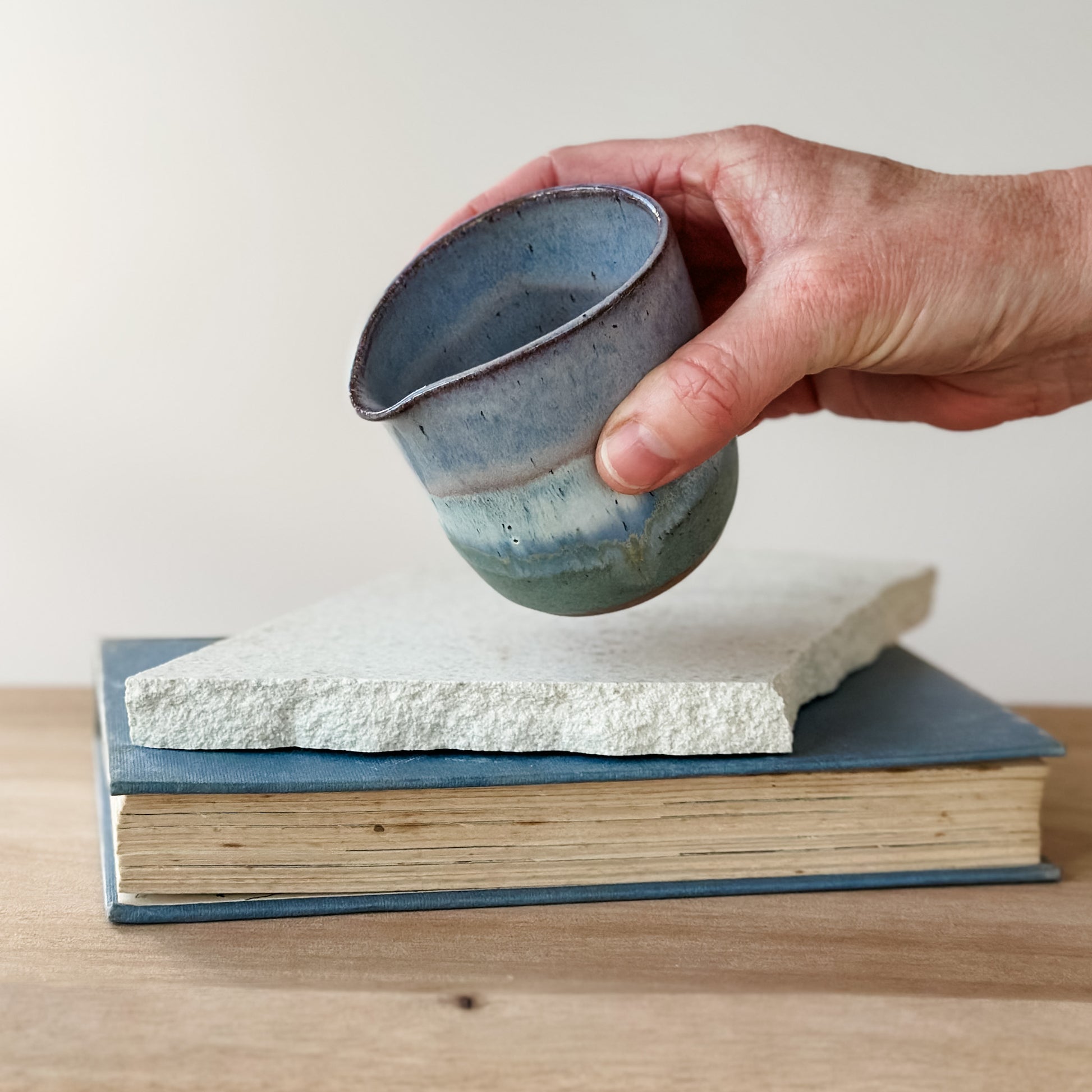 Hand holding a blue ceramic creamer jug over a stack of books on a wooden surface.