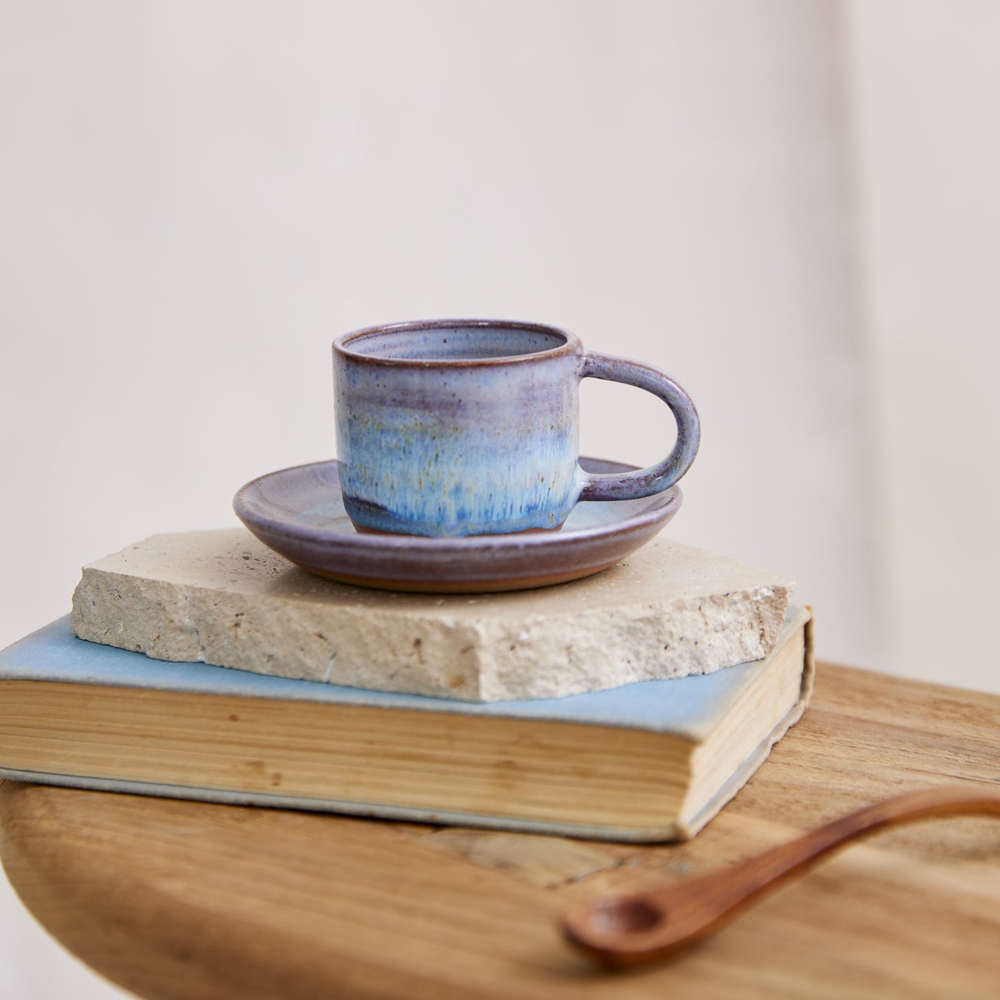 A front view of the Coastal Handmade Ceramic Espresso Cup and saucer. The handmade mug and saucer have a lavender, blue and white glaze. The handmade espresso mug and saucer are on a tile, book and wooden table in a coastal setting.