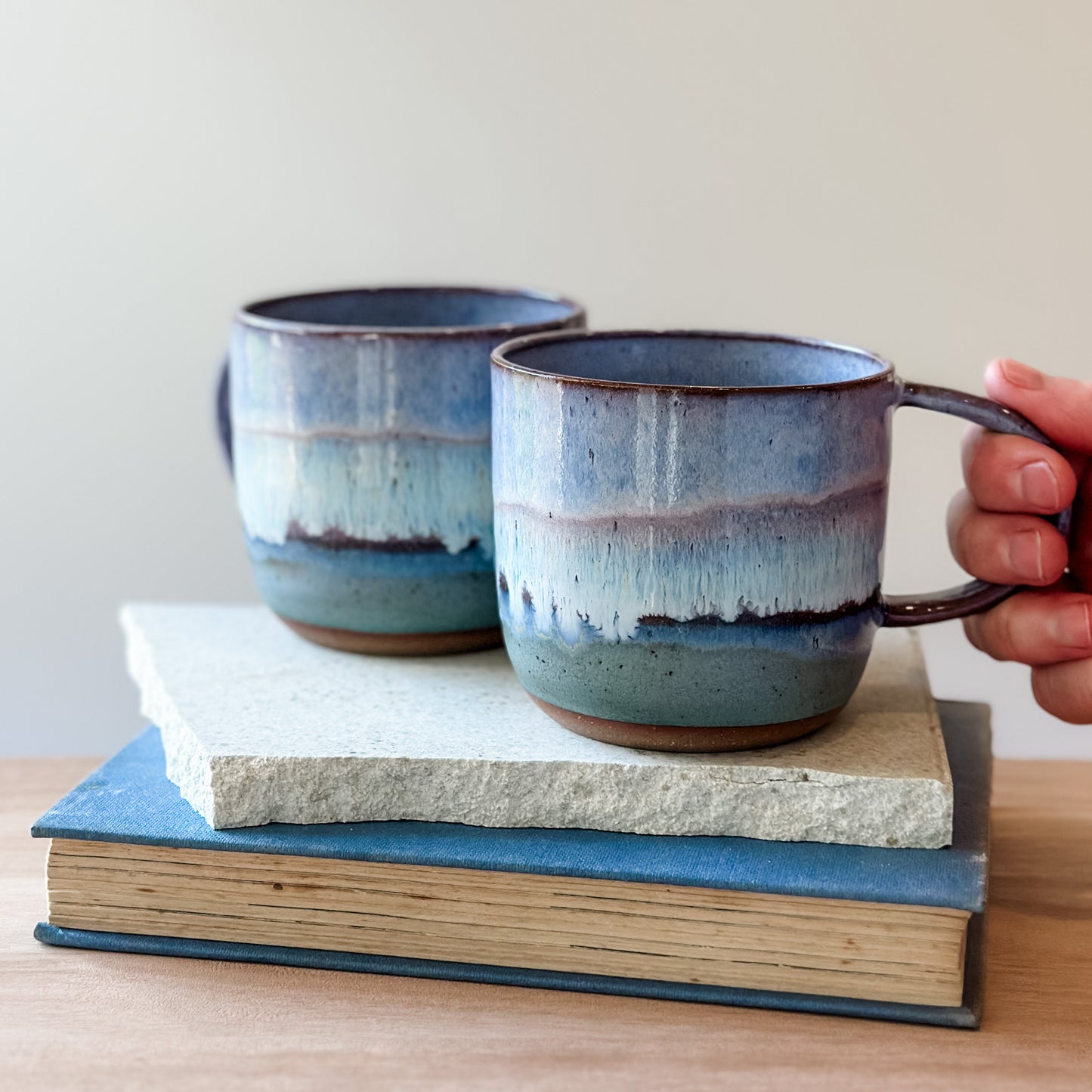Two purple and blue ceramic mugs on top of books, with a hand holding one mug.