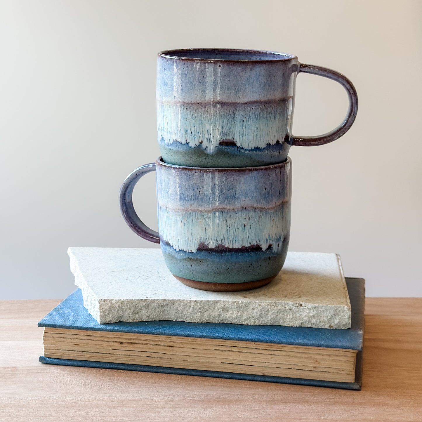 Two stacked ceramic mugs with a textured finish on top of a tile and a book.