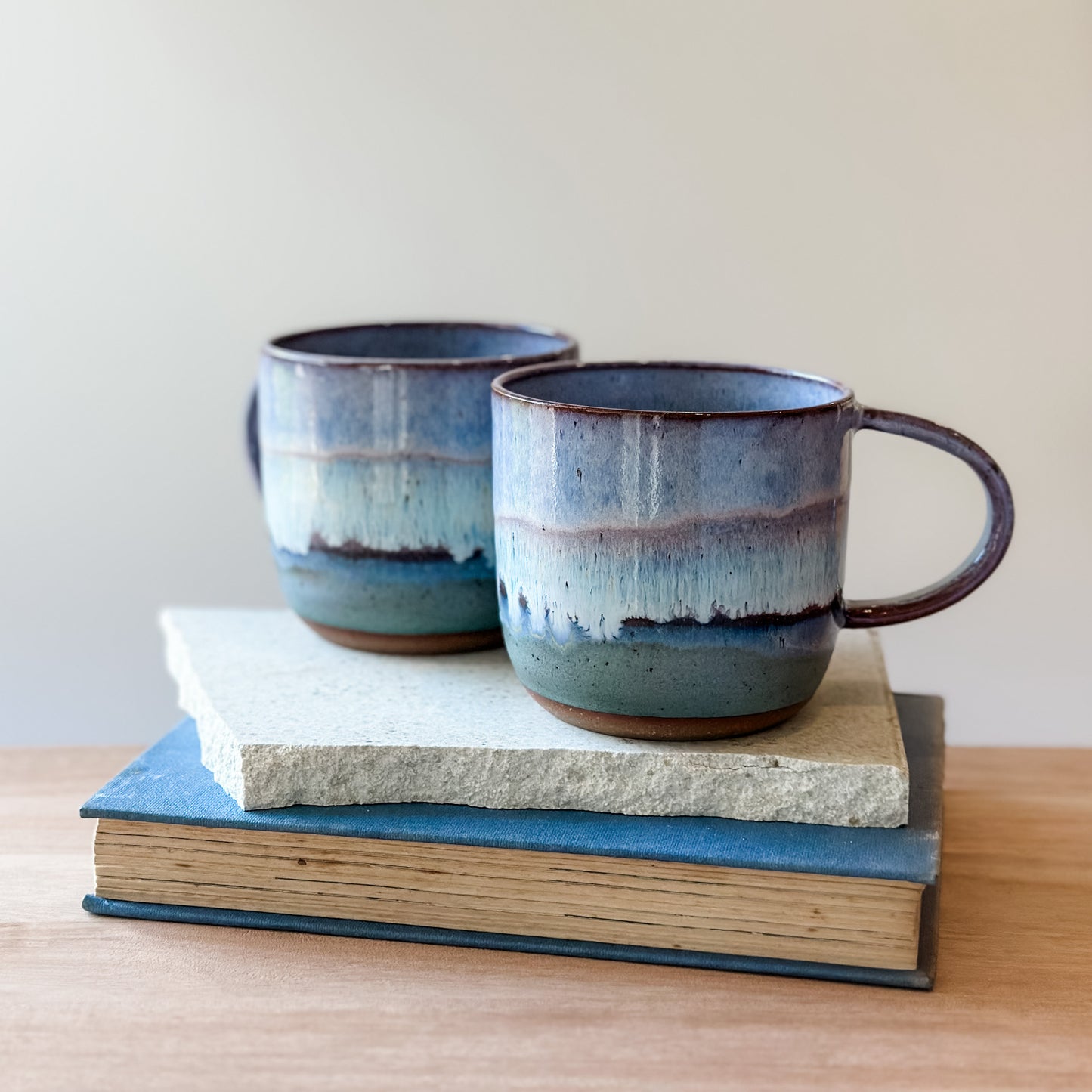 Two ceramic mugs with blue and purple glaze on top of a stack of books.