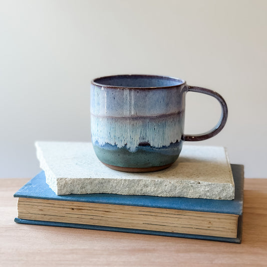 Ceramic mug with purple and blue glaze on top of a book and a tile