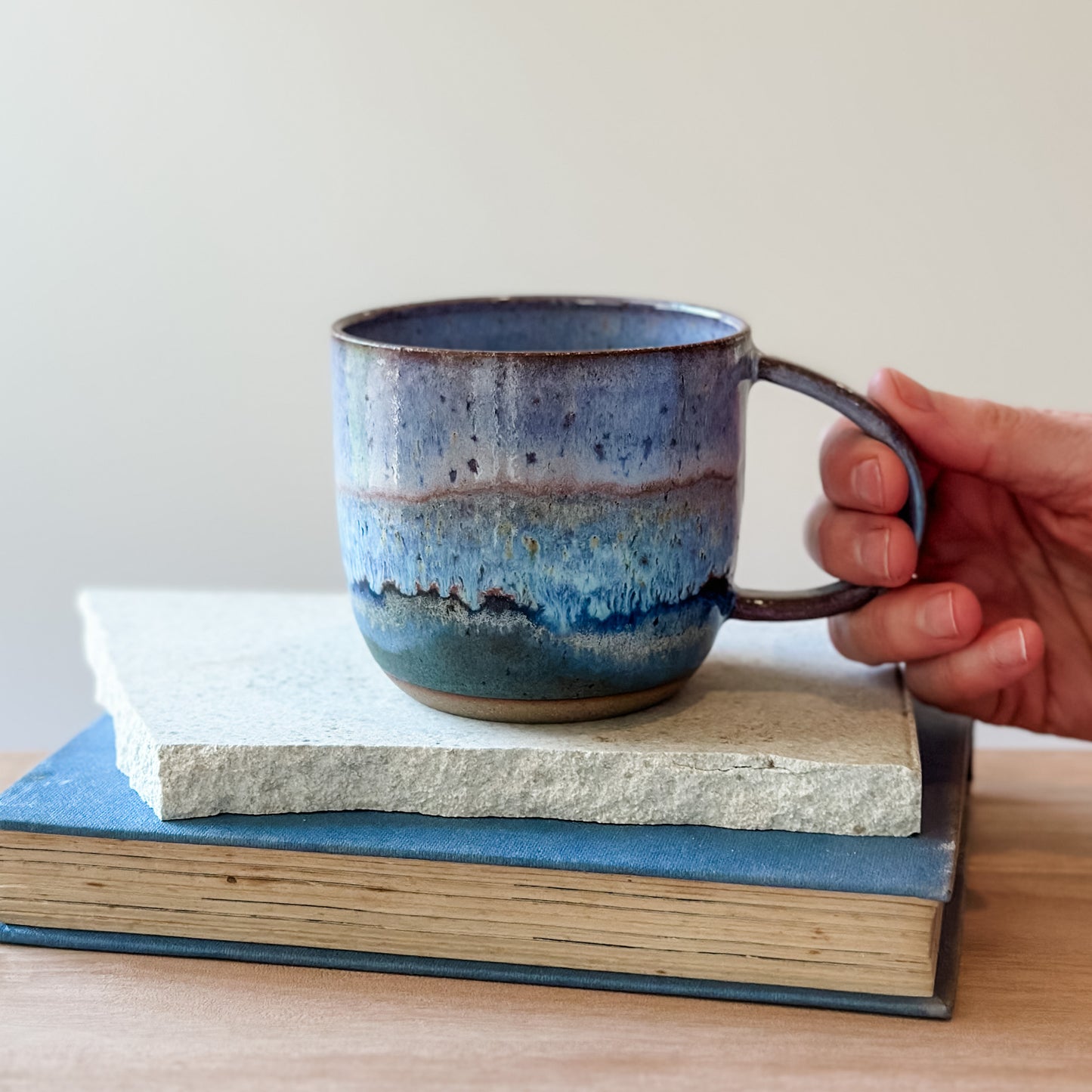 Hand holding a blue ceramic mug over a stack of books on a wooden surface.