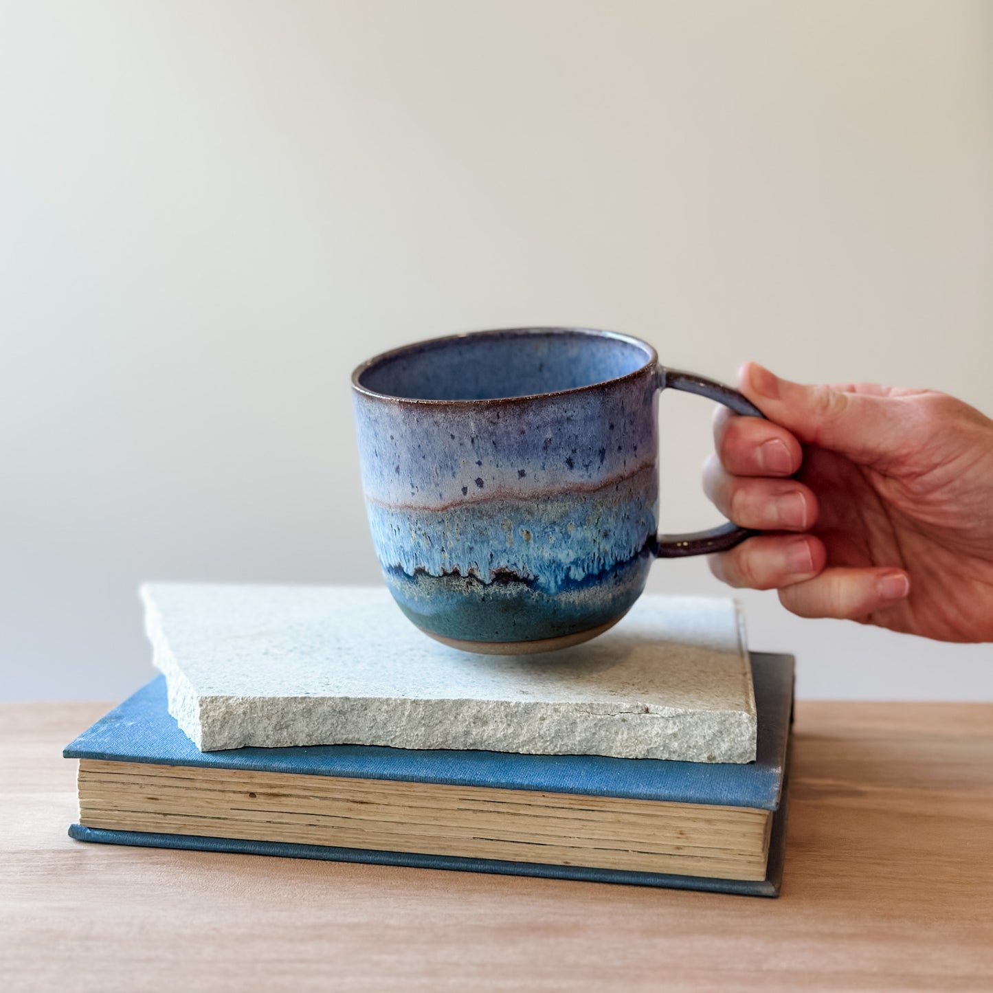Hand holding a blue ceramic mug over a book on a wooden surface.