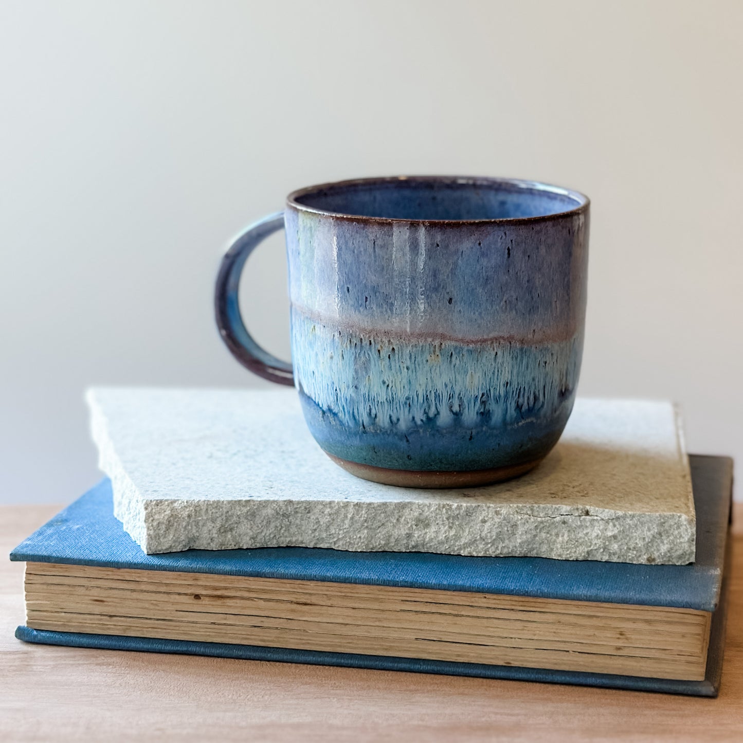 A purple and blue ceramic mug is on a stack of books and a table.