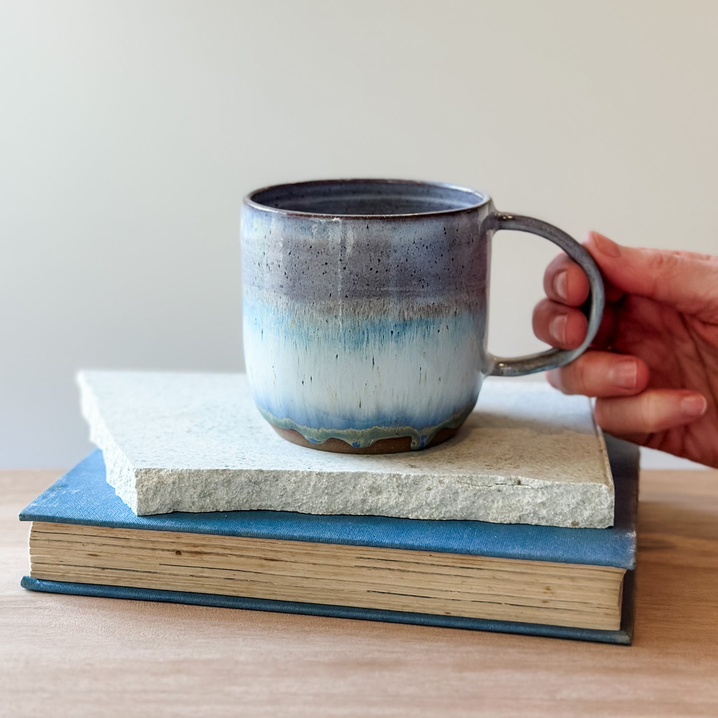 Hand holding a ceramic mug over a book and a tile on a wooden surface.