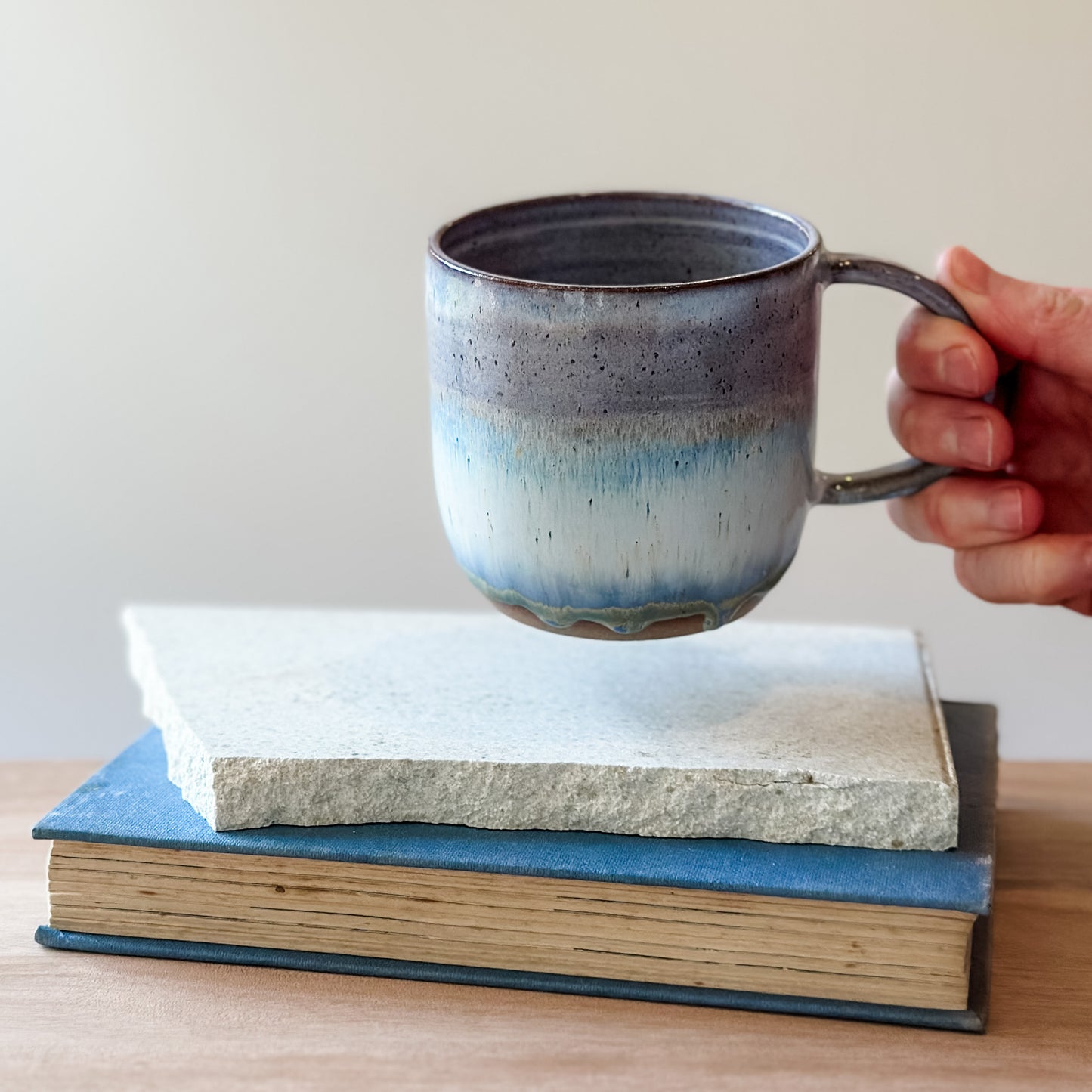 Hand holding a ceramic mug over a tile and a book on a wooden surface.