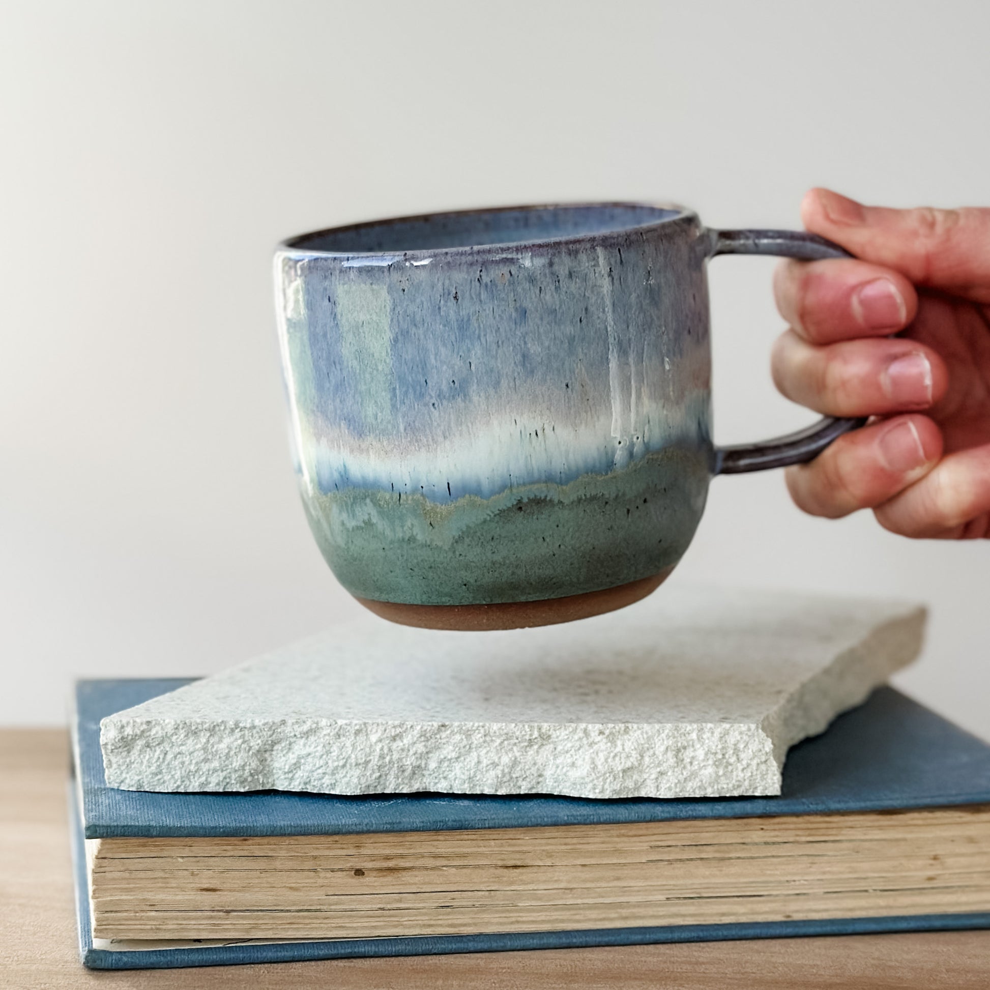 Hand holding a blue ceramic mug over a stack of books on a light surface.