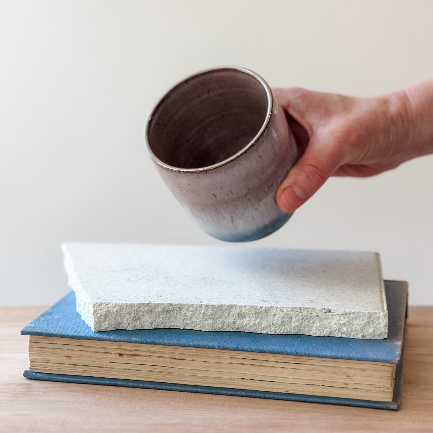 Hand holding a ceramic cup over a tile and a book on a wooden surface.