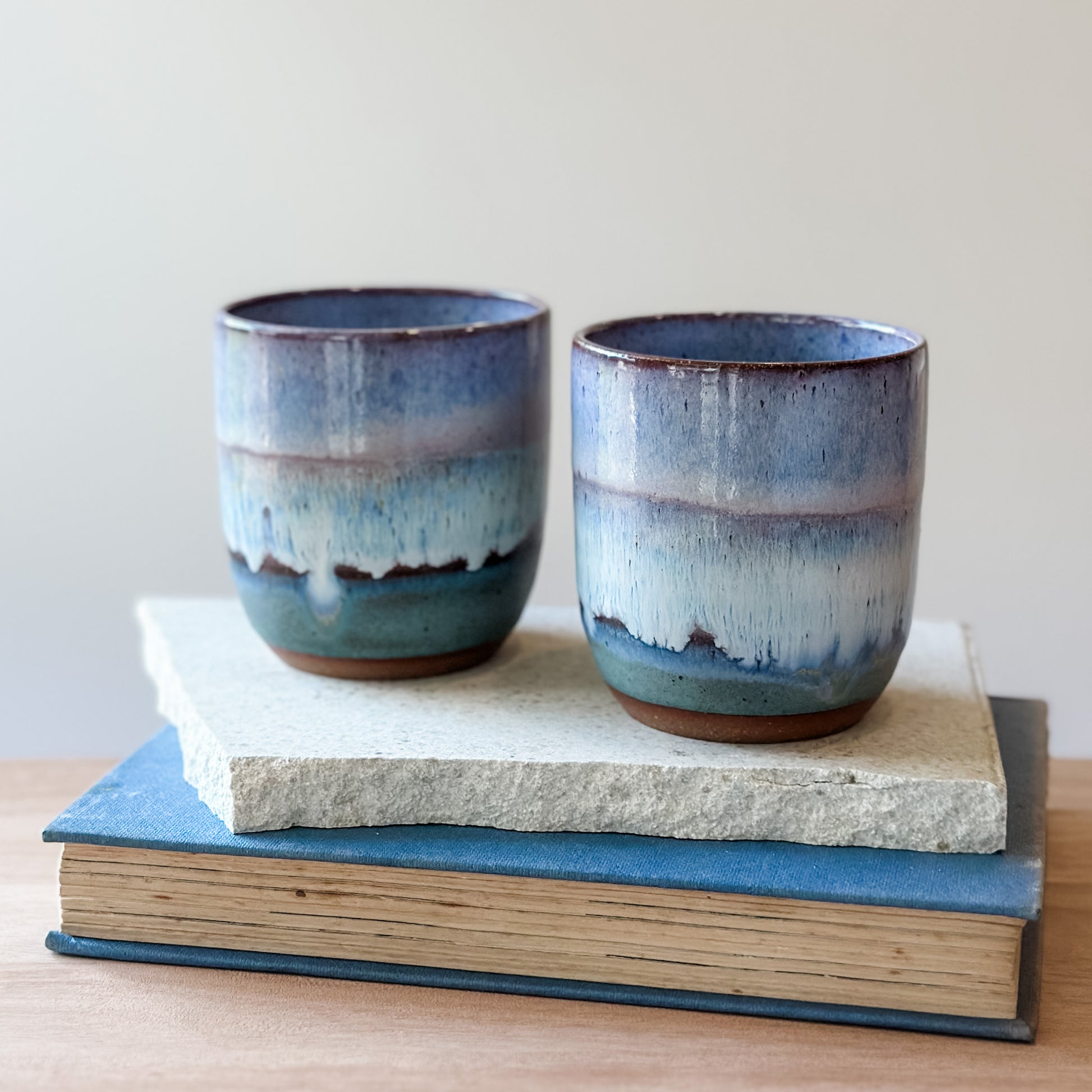 Two ceramic cups with blue and purple glaze on top of a tile and a book.