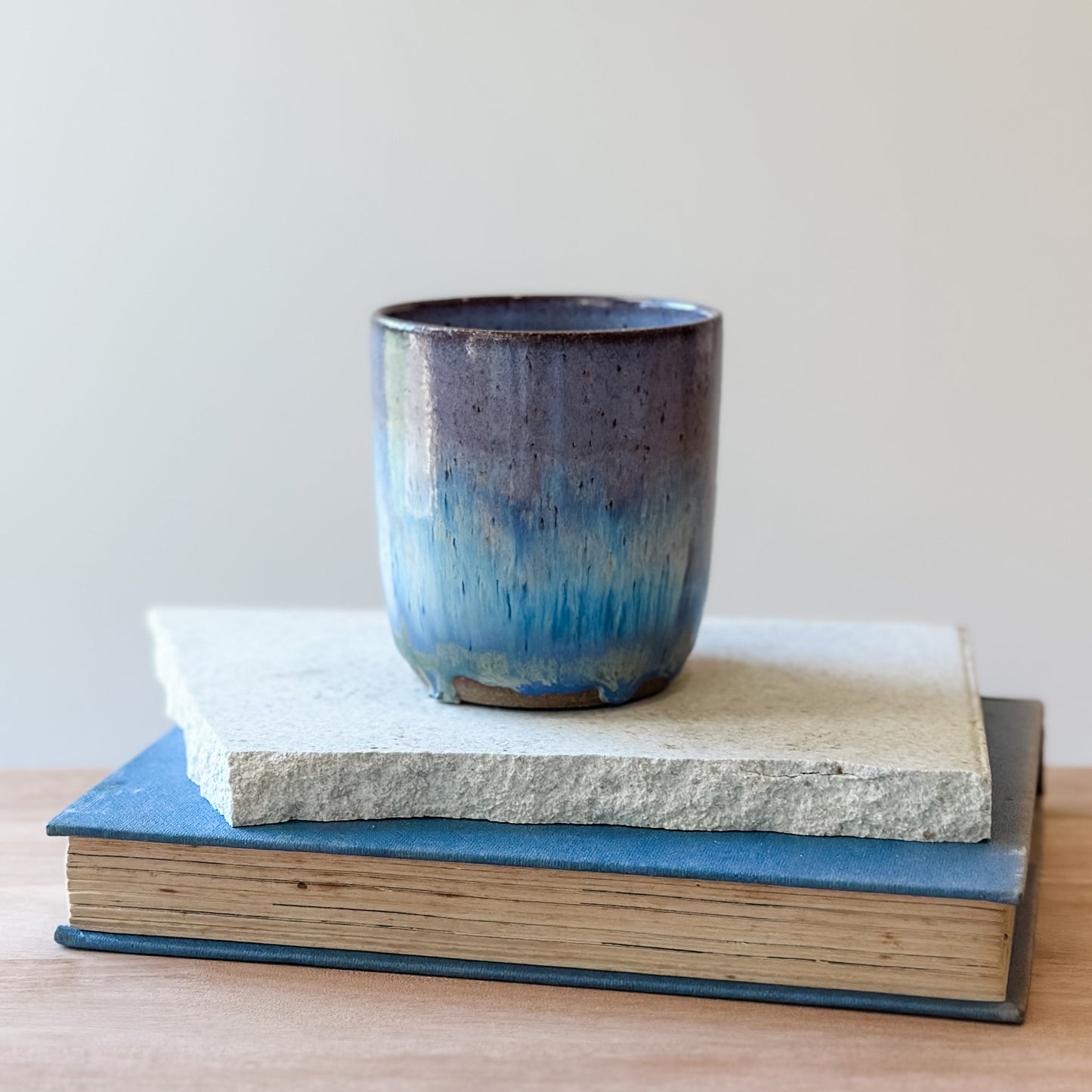A purple and blue ceramic cup on top of a tile and a book with a plain background