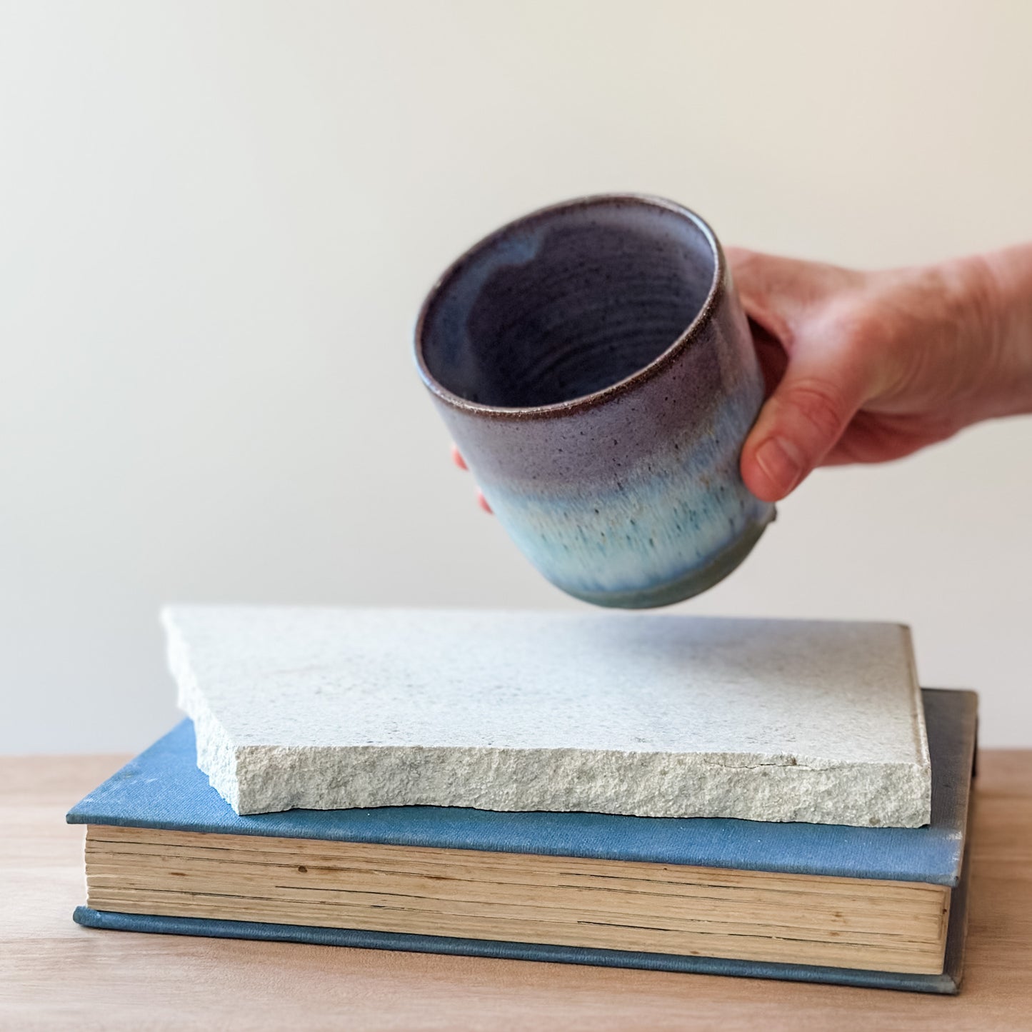 Hand holding a ceramic cup over two stacked books on a light surface.