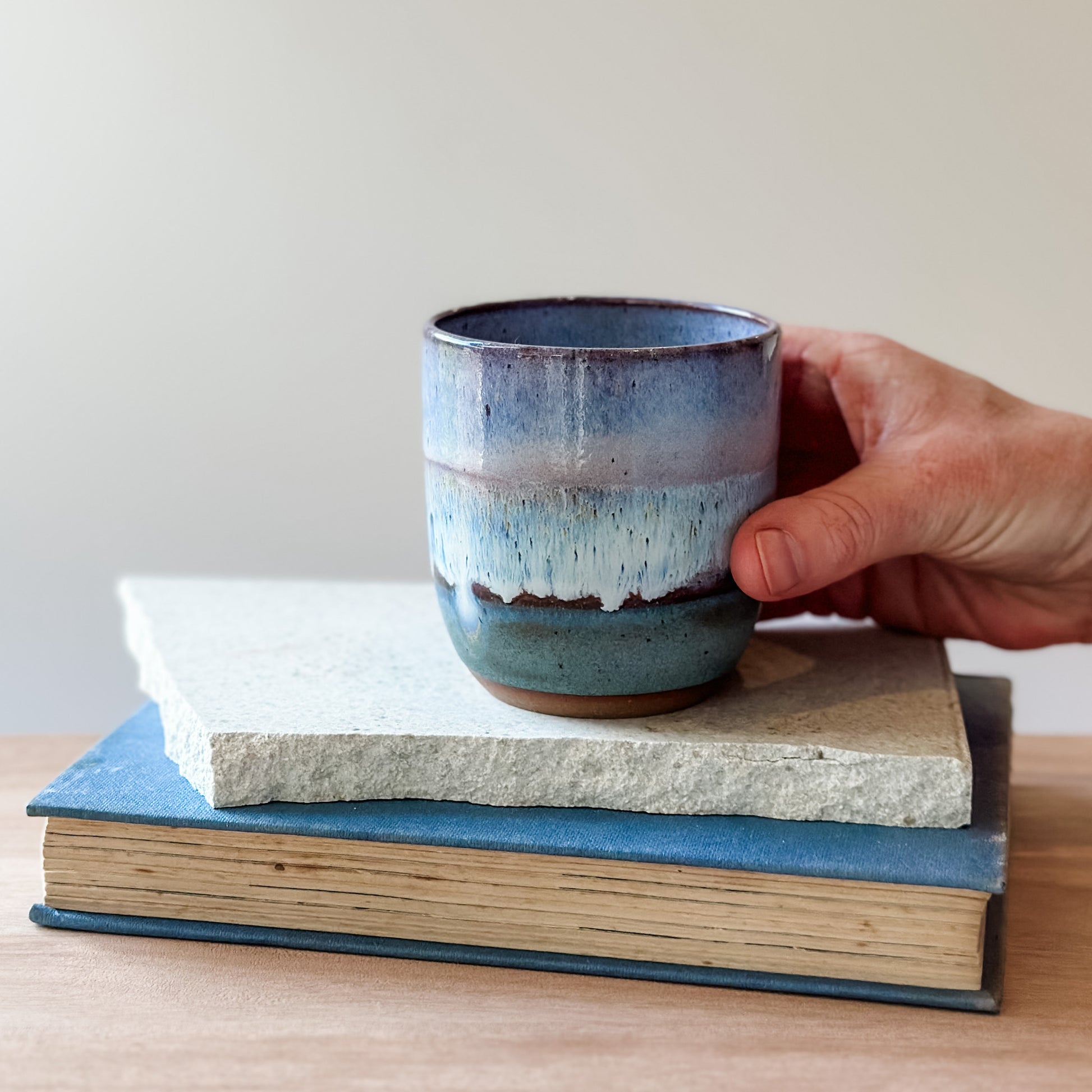 Hand holding a purple and blue ceramic cup over a stack of books on a wooden surface.