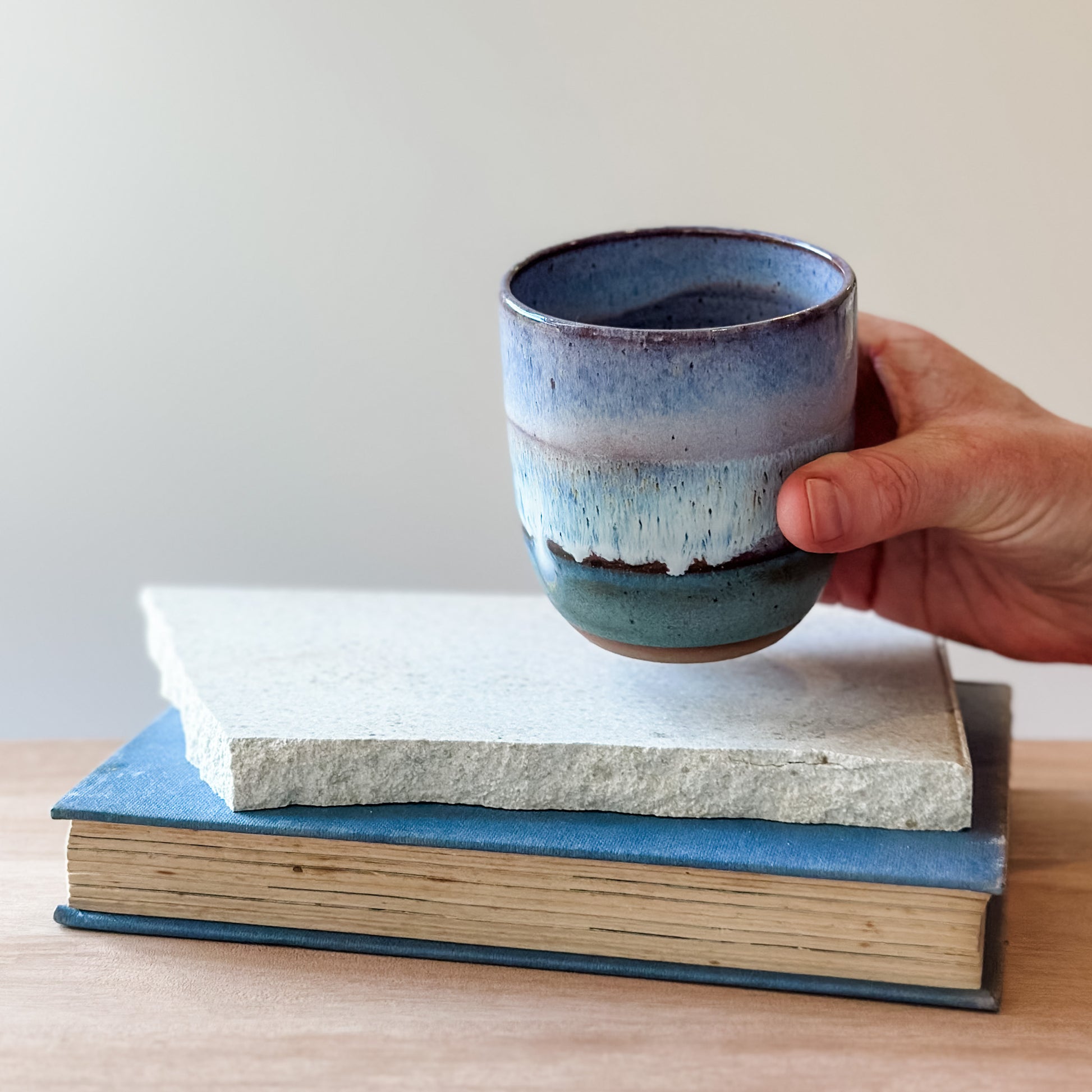 Hand holding a purple and blue ceramic cup over a tile and a book on a wooden surface.