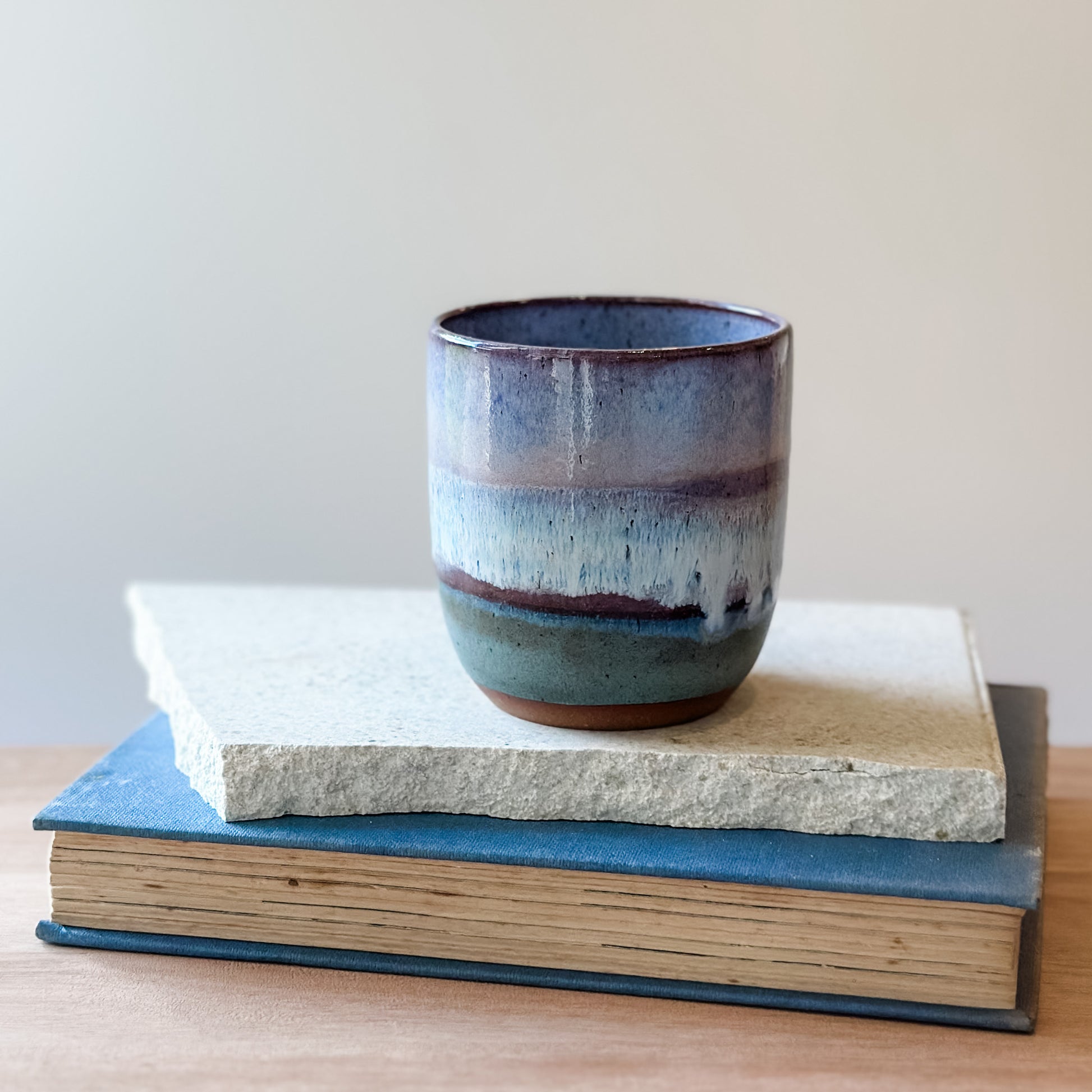 Ceramic cup with purple and blue glaze on top of a book and a tile on a wooden surface.