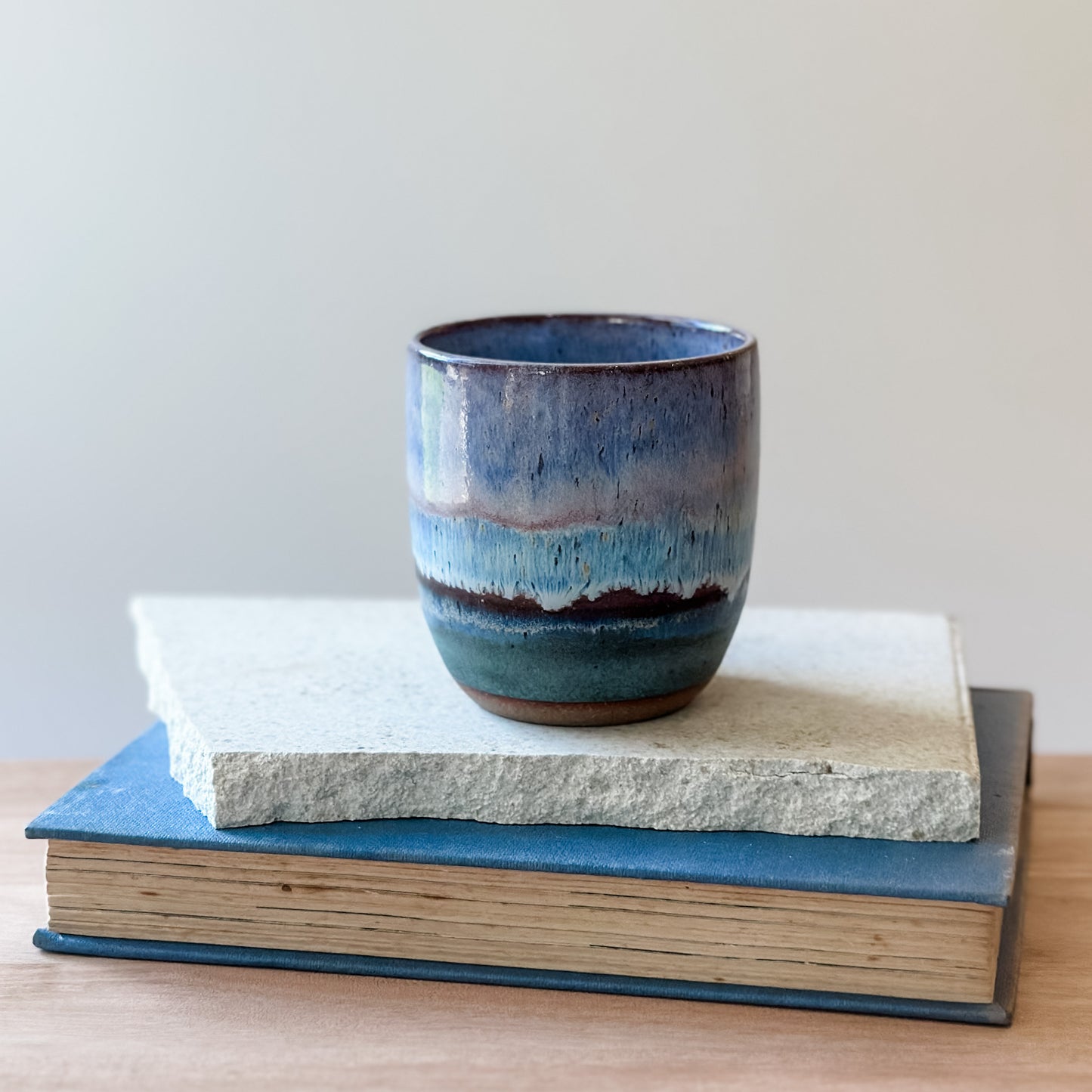 Ceramic mug with blue and purple glaze on top of a book and tile.