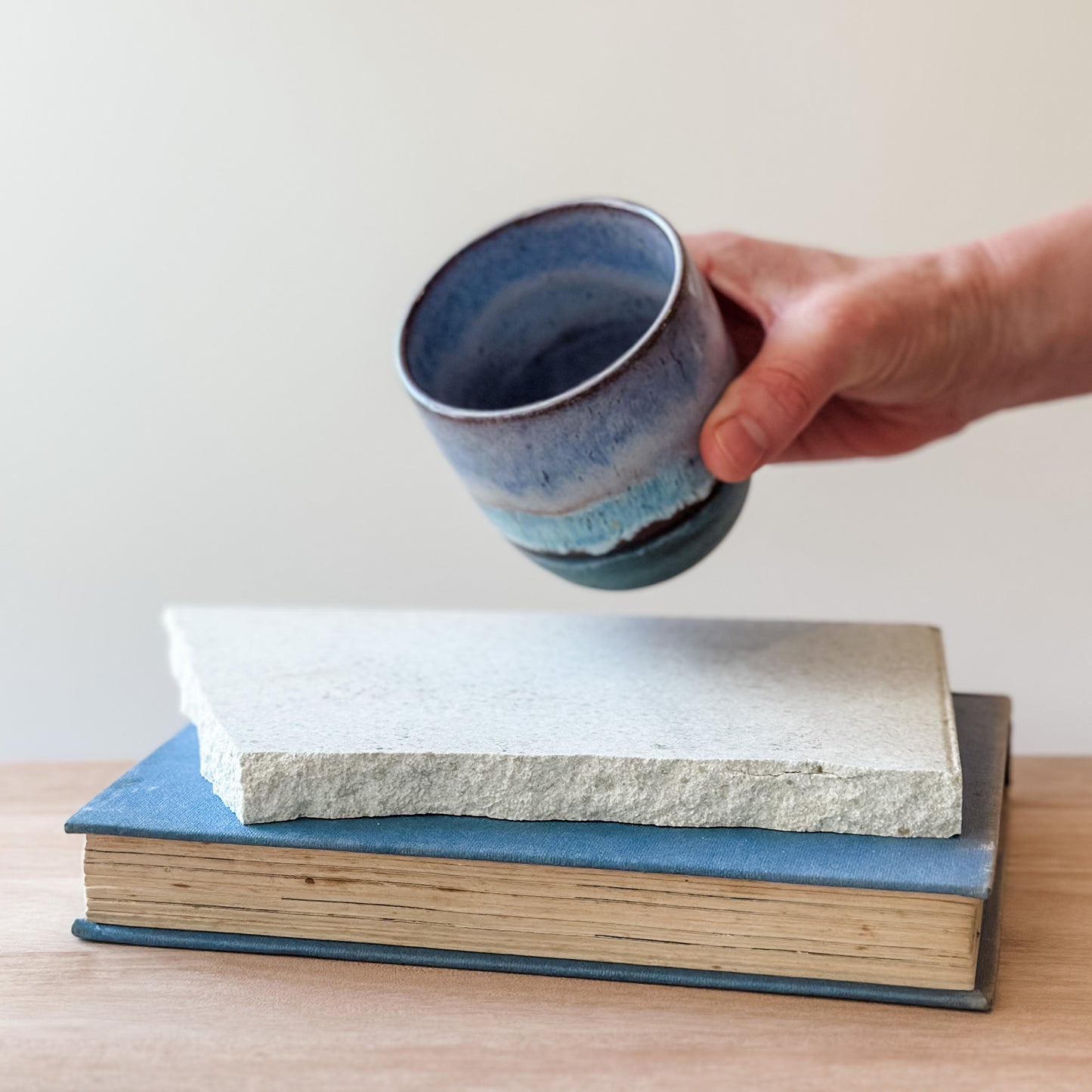Hand holding a blue ceramic cup over a tile and a book on a wooden surface.