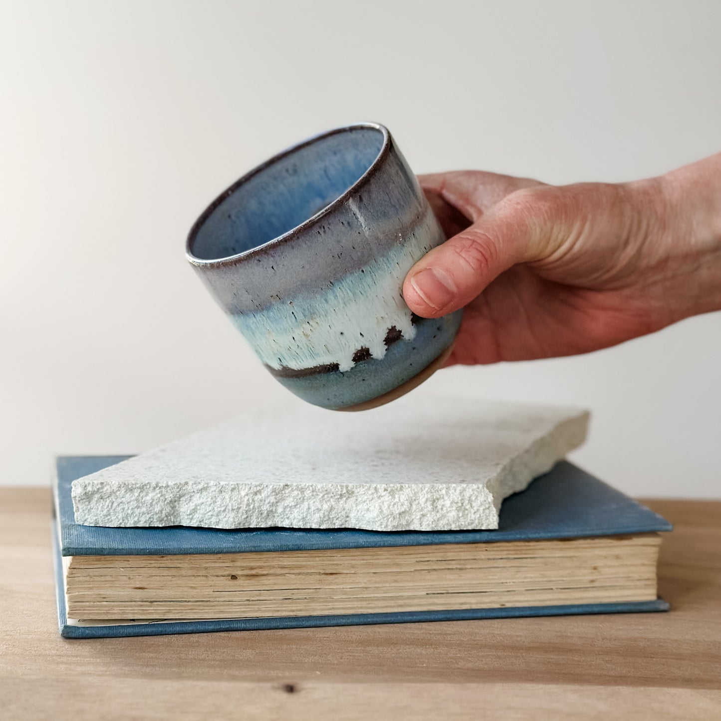 Hand holding a ceramic cup over a stack of books on a wooden surface.