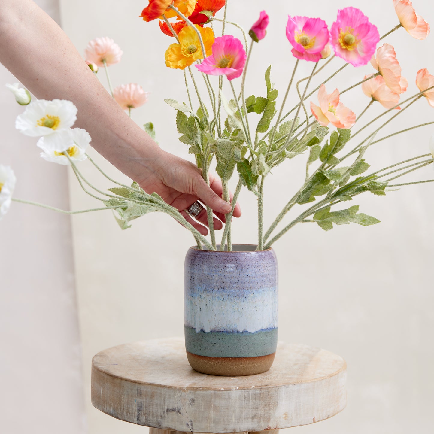 A front view of the Coastal Handmade Ceramic Vase in a wide style. This hand-painted vase is glazed in lavender, white and blue glaze. The ceramic vase is sitting on a wooden stool in a coastal-styled setting. The vase is displaying colourful flowers which are being arranged by hand.