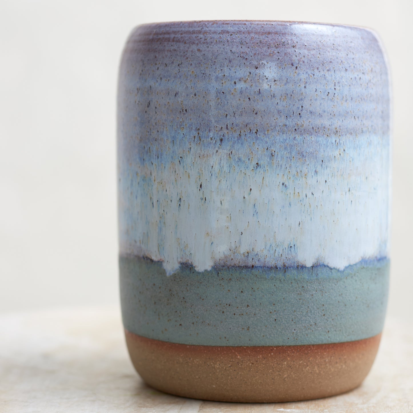 A close-up view of the Coastal Handmade Ceramic Vase in a wide style. This hand-painted vase is glazed in lavender, white and blue glaze. The ceramic vase is sitting on a wooden stool in a coastal-styled setting.
