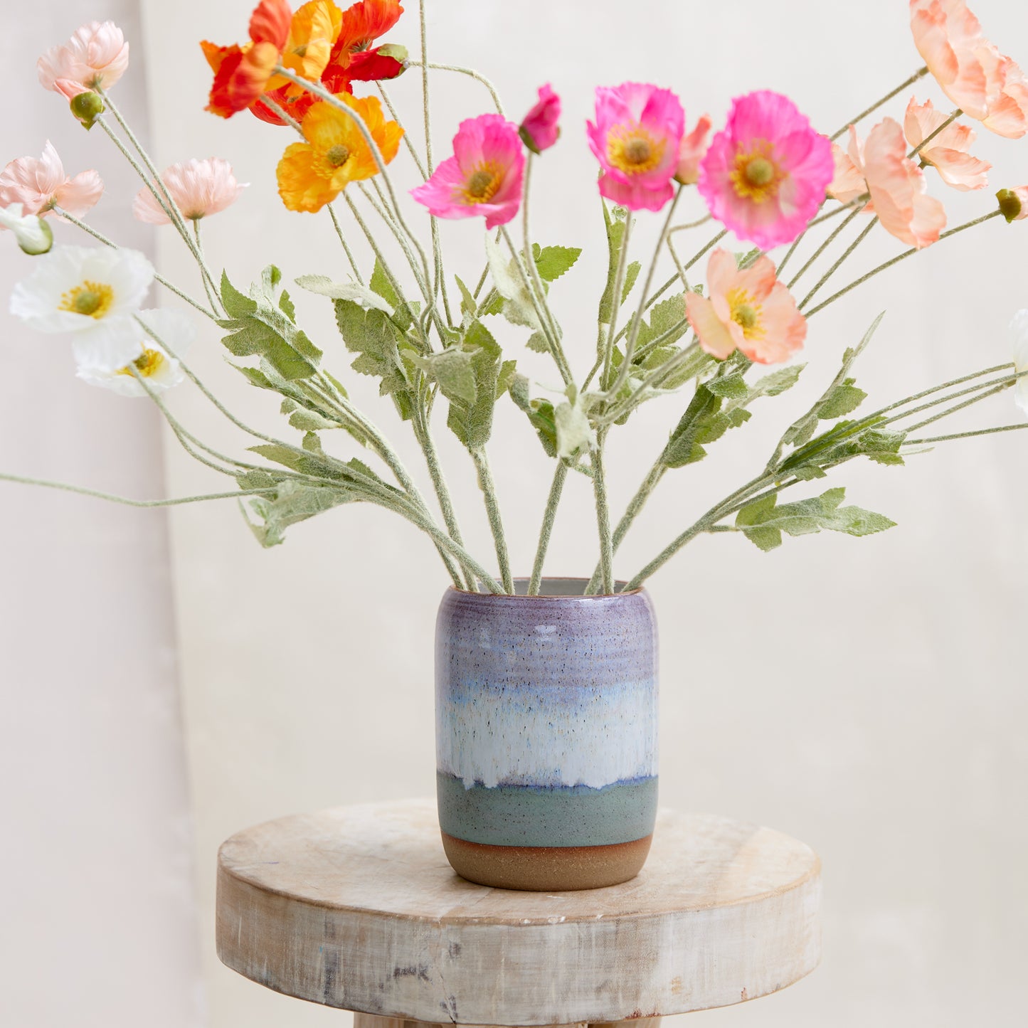A front view of the Coastal Handmade Ceramic Vase in a wide style. This hand-painted vase is glazed in lavender, white and blue glaze. The ceramic vase is sitting on a wooden stool in a coastal-styled setting. The vase is displaying colourful flowers.