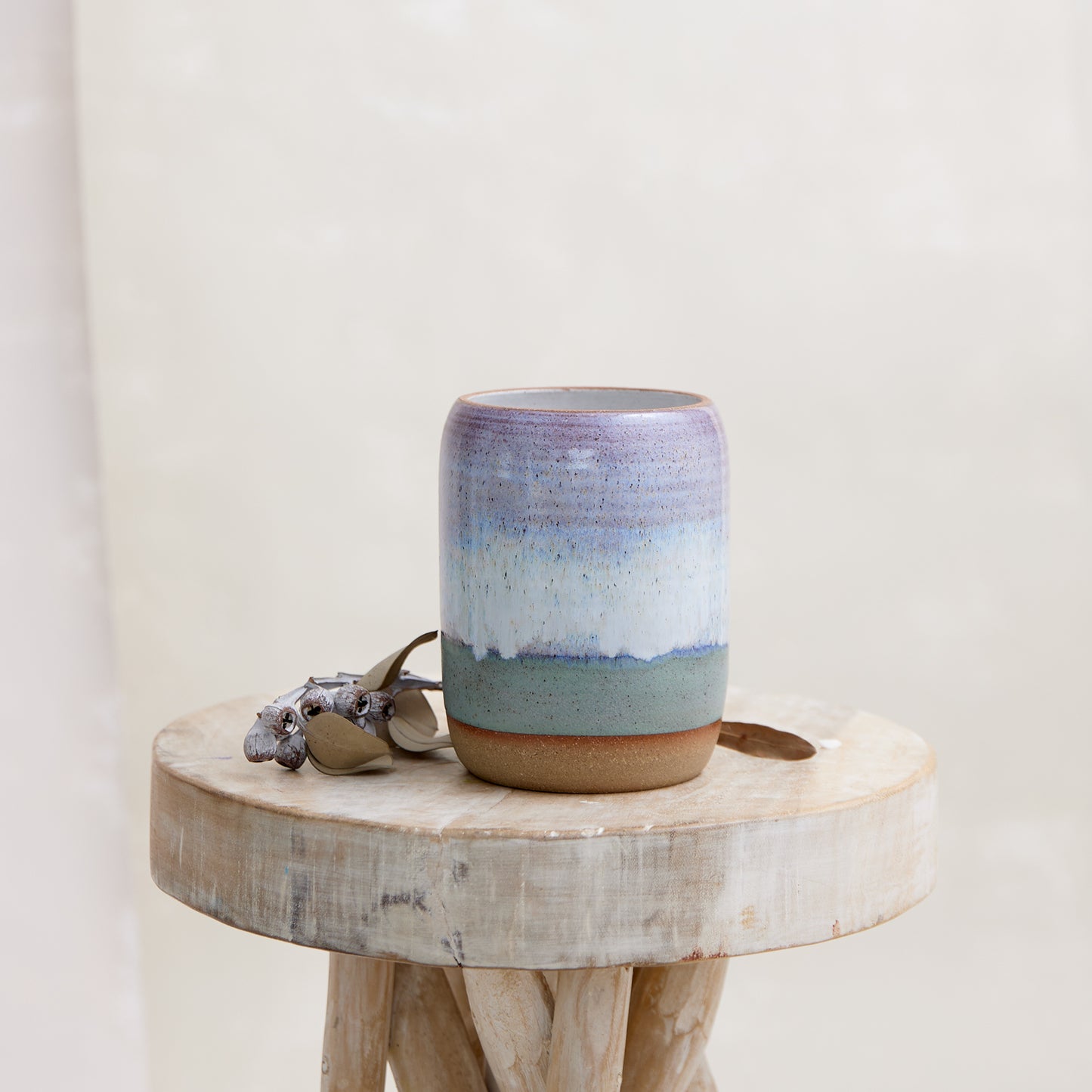 A front view of the Coastal Handmade Ceramic Vase in a wide style. This hand-painted vase is glazed in lavender, white and blue glaze. The ceramic vase is sitting on a wooden stool in a coastal-styled setting.