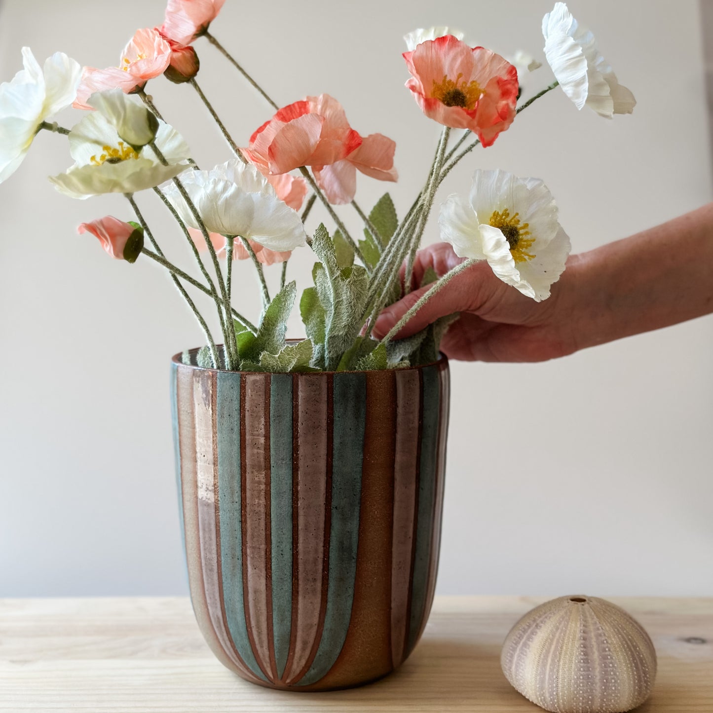 A large handmade ceramic bouquet vase sits on a table. The ceramic vase is hand-painted in blush pink and denim blue glaze. A hand is arranging flowers in the bouquet vase.