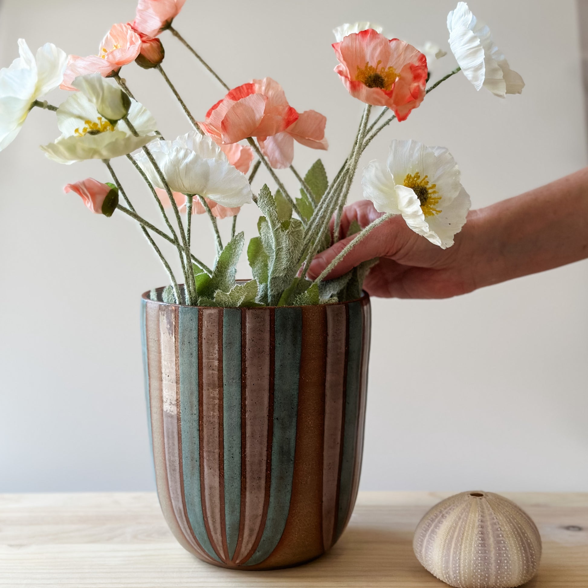 A large handmade ceramic bouquet vase sits on a table. The ceramic vase is hand-painted in blush pink and denim blue glaze. A hand is arranging flowers in the bouquet vase.