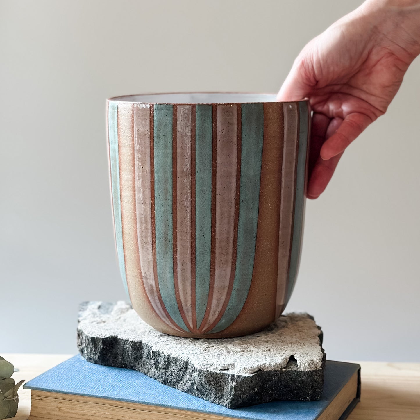 A large handmade ceramic bouquet vase sits on a table. The ceramic vase is hand-painted in blush pink and denim blue glaze. A hand is placing the handmade ceramic vase onto a tile on top of a table.