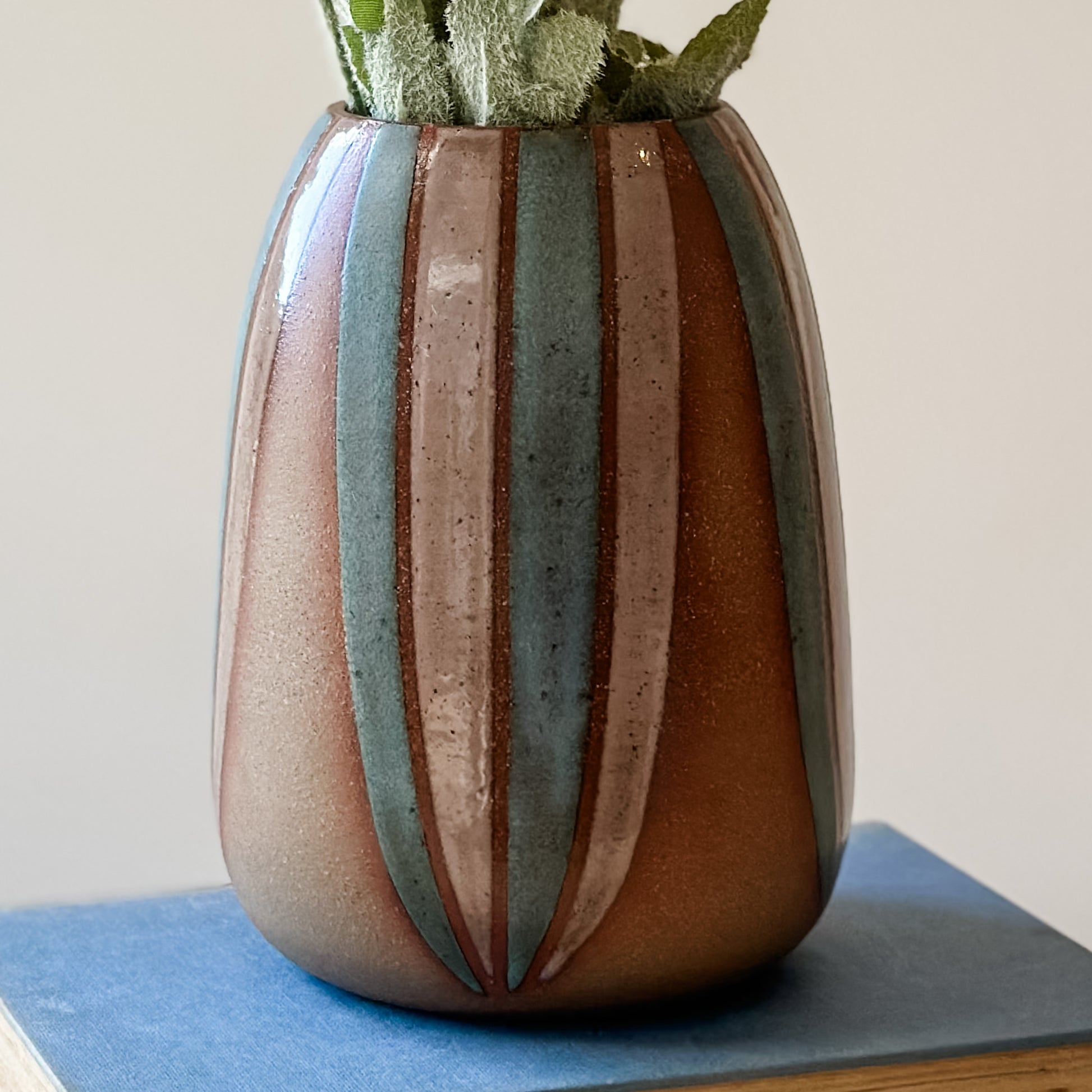 A close-up view of a handmade ceramic vase, hand-painted in blush pink and blue. The bellied ceramic vase sits on a book.