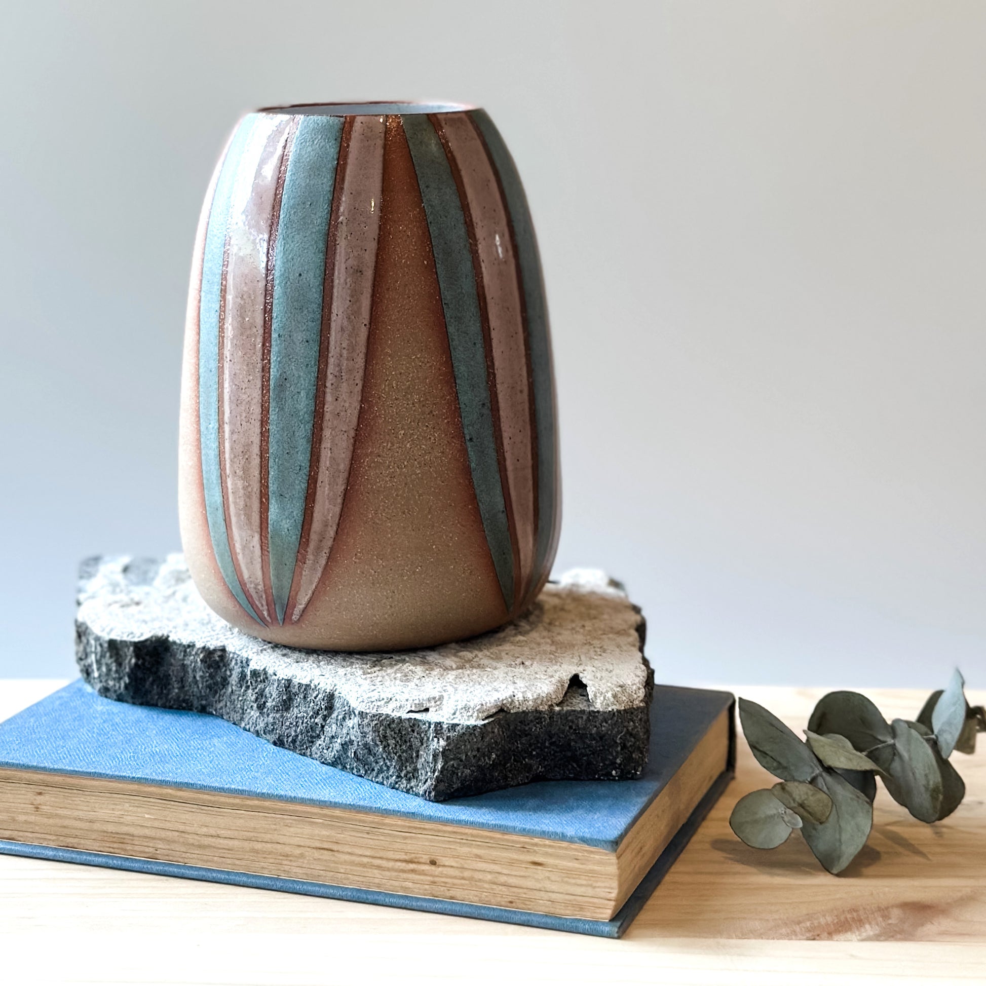 A side view of a handmade ceramic vase, hand-painted in blush pink and blue. The bellied ceramic vase is sitting on a tile and a book on top of a table.