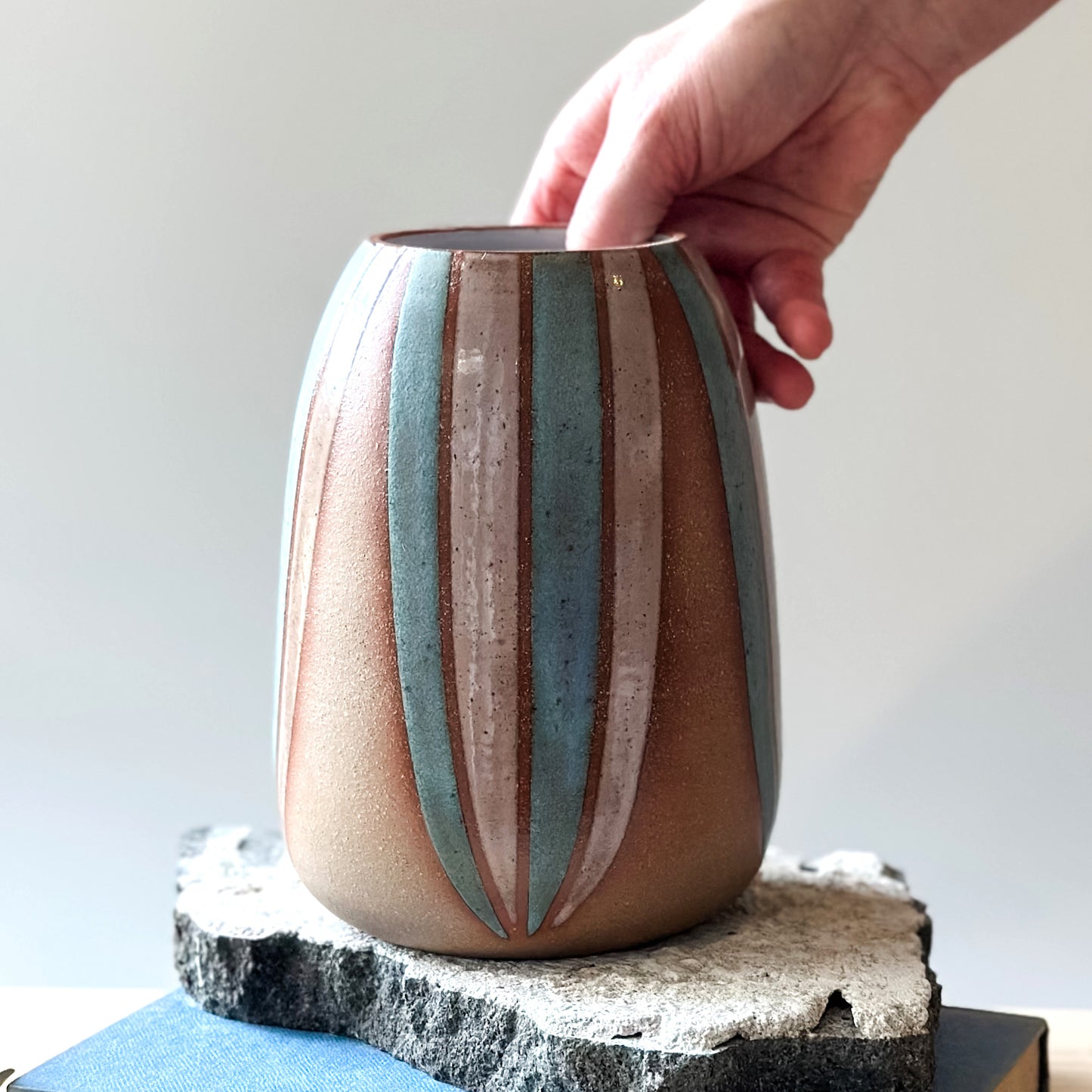 A front view of a handmade ceramic vase, hand-painted in blush pink and blue. The bellied ceramic vase is being placed by hand on a tile on top of a table.