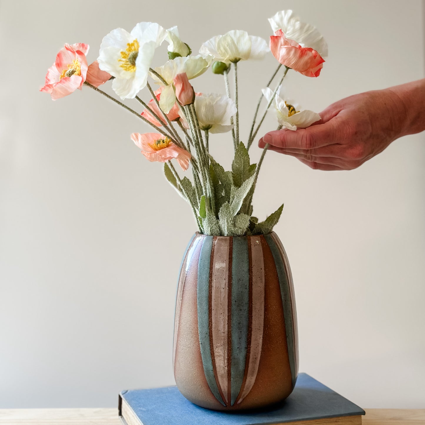 A front view of a handmade ceramic vase, hand-painted in blush pink and blue. Flowers are being arranged by hand in the vase. The bellied ceramic vase sits on a book on top of a table.