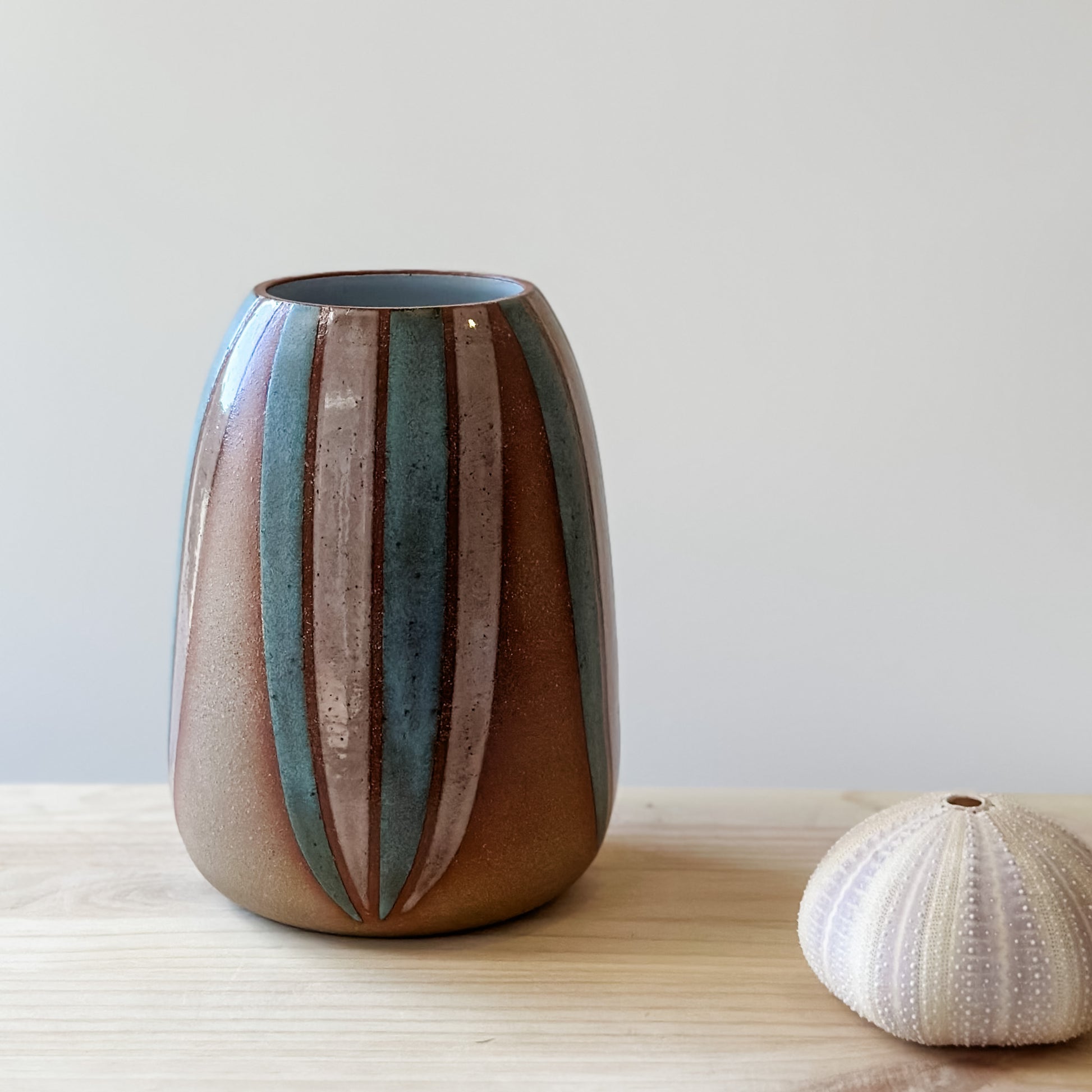 A front view of a handmade ceramic vase, hand-painted in blush pink and blue. The bellied ceramic vase sits on a wooden table with a seashell.