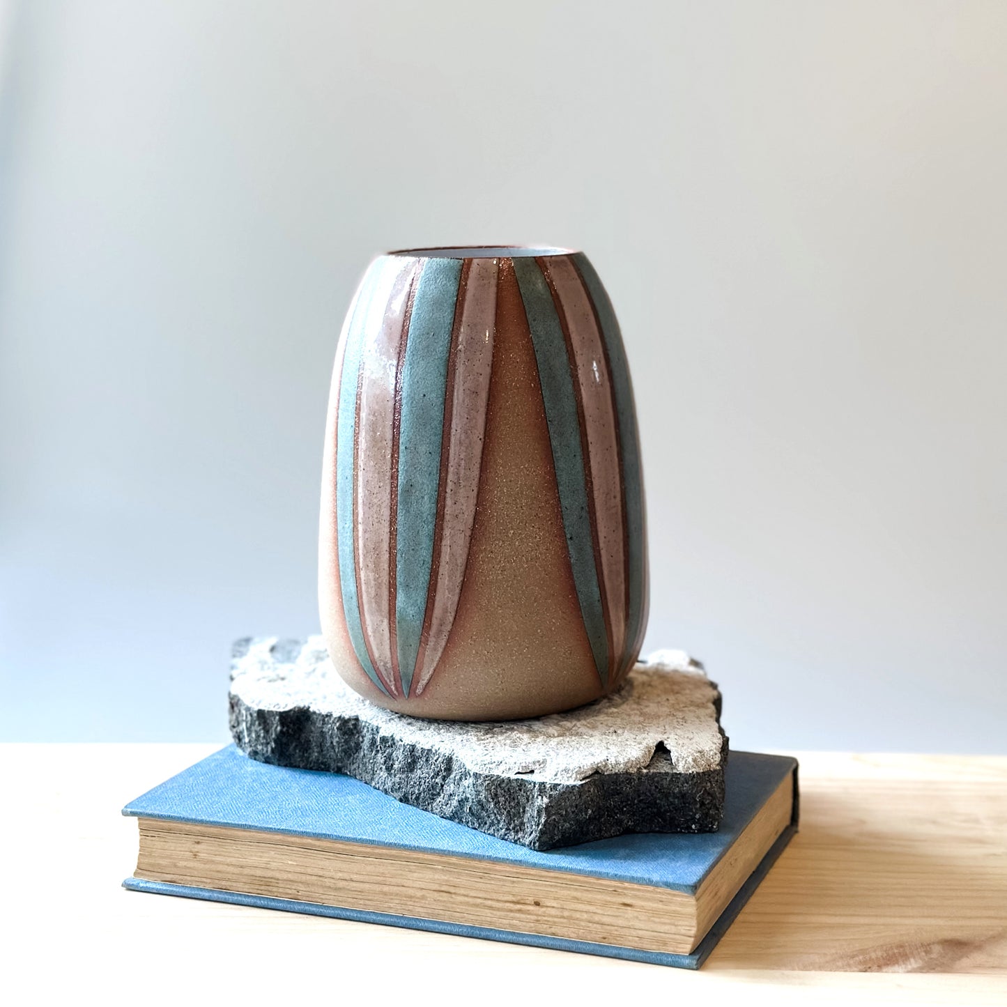A side view of a handmade ceramic vase, hand-painted in blush pink and blue stripes. The bellied ceramic vase sits on a tile and a book on top of a wooden table.