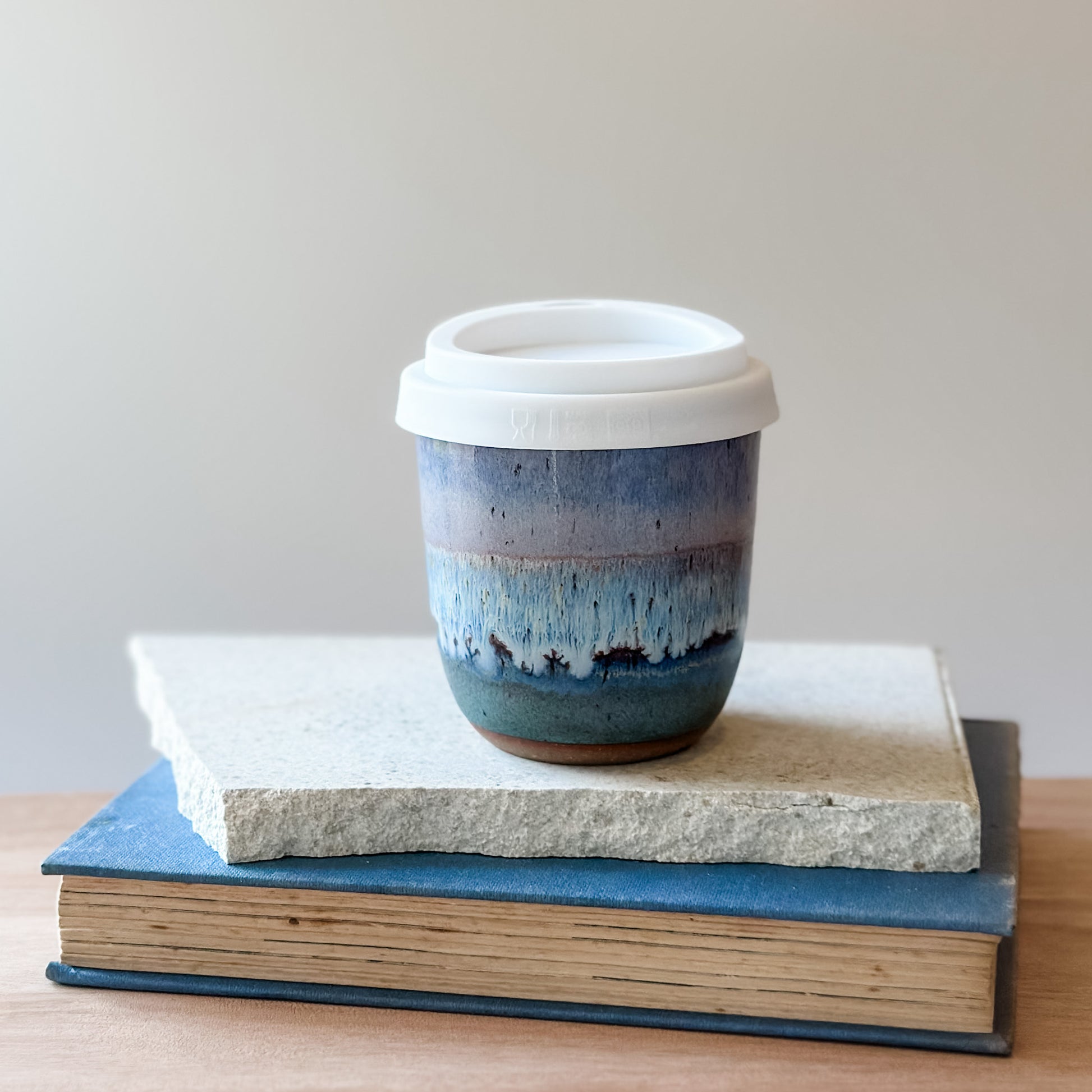Purple, white, and blue ceramic cup with a white lid on top of a tile and a book