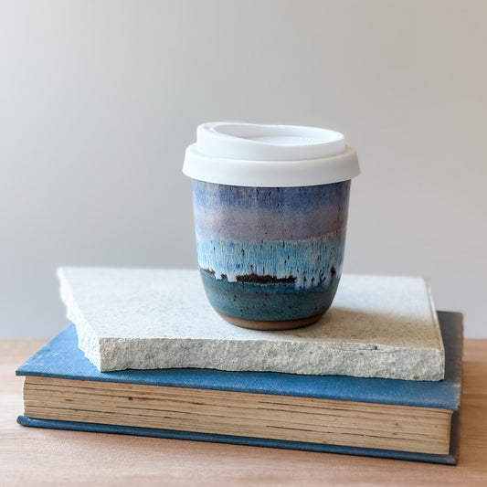 Purple and blue ceramic coffee cup with a white lid on top of a tile and a book