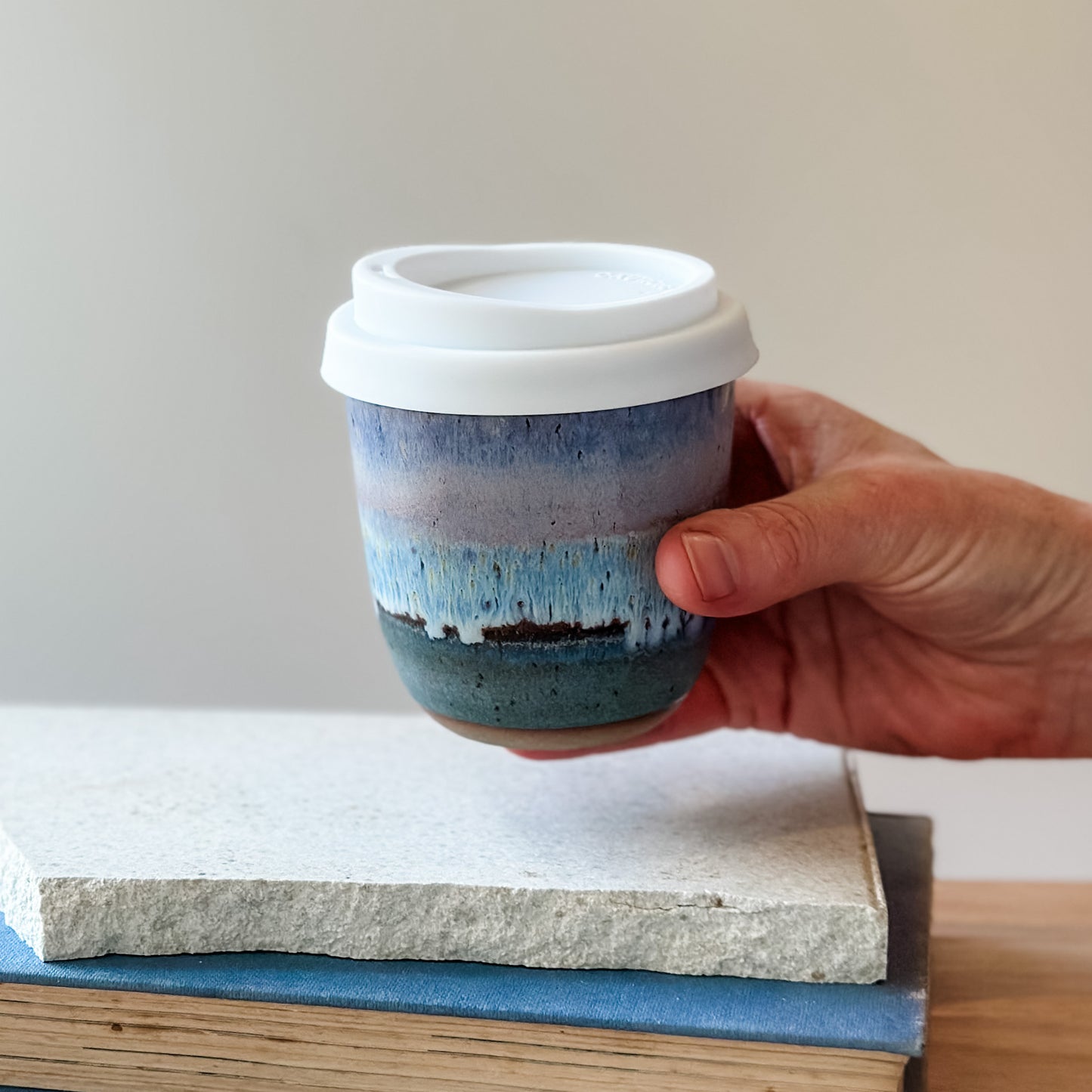 Hand holding a ceramic keep cup with a blue and white design on a wooden surface.