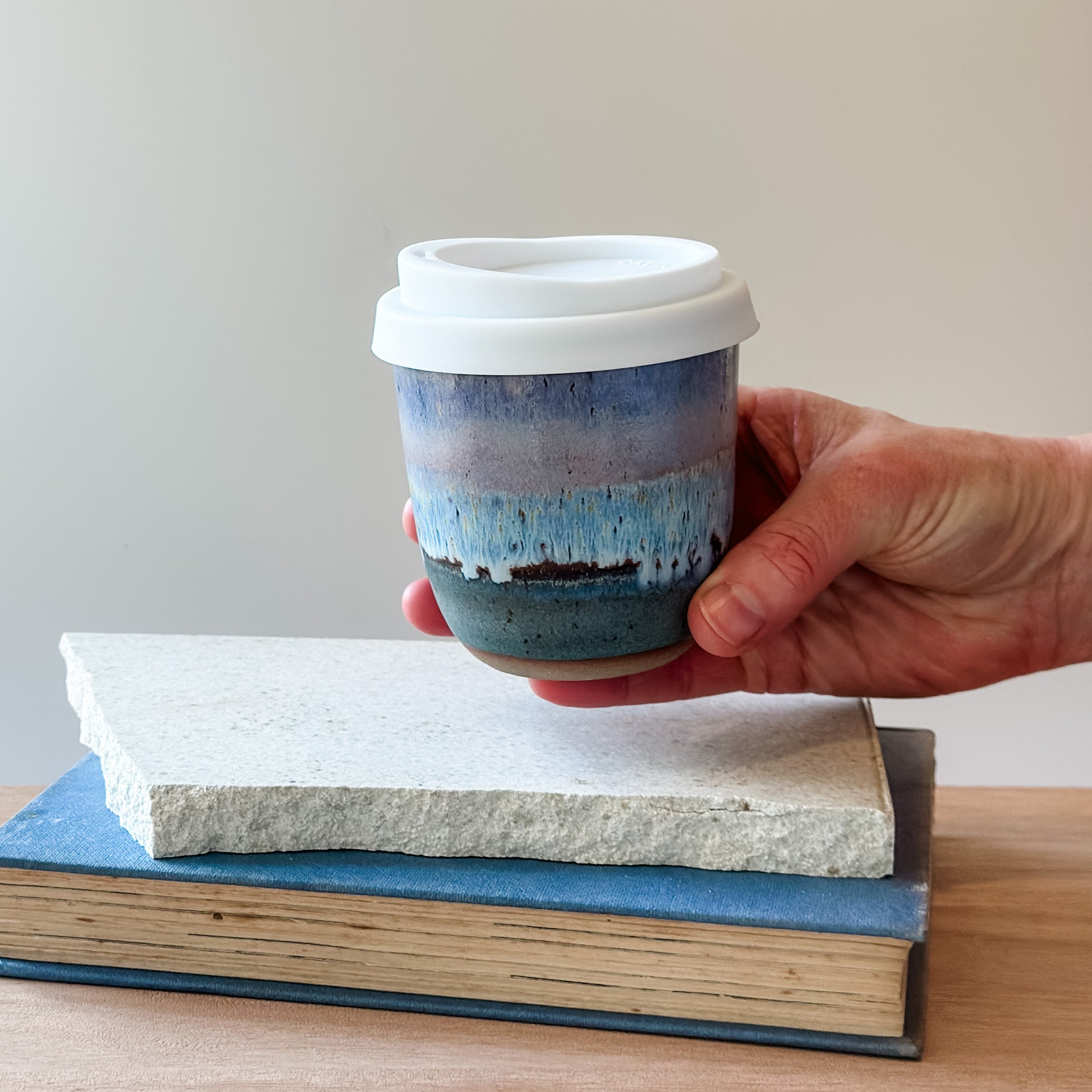 Hand holding a purple and blue ceramic cup with a white lid over a stack of books on a wooden surface.