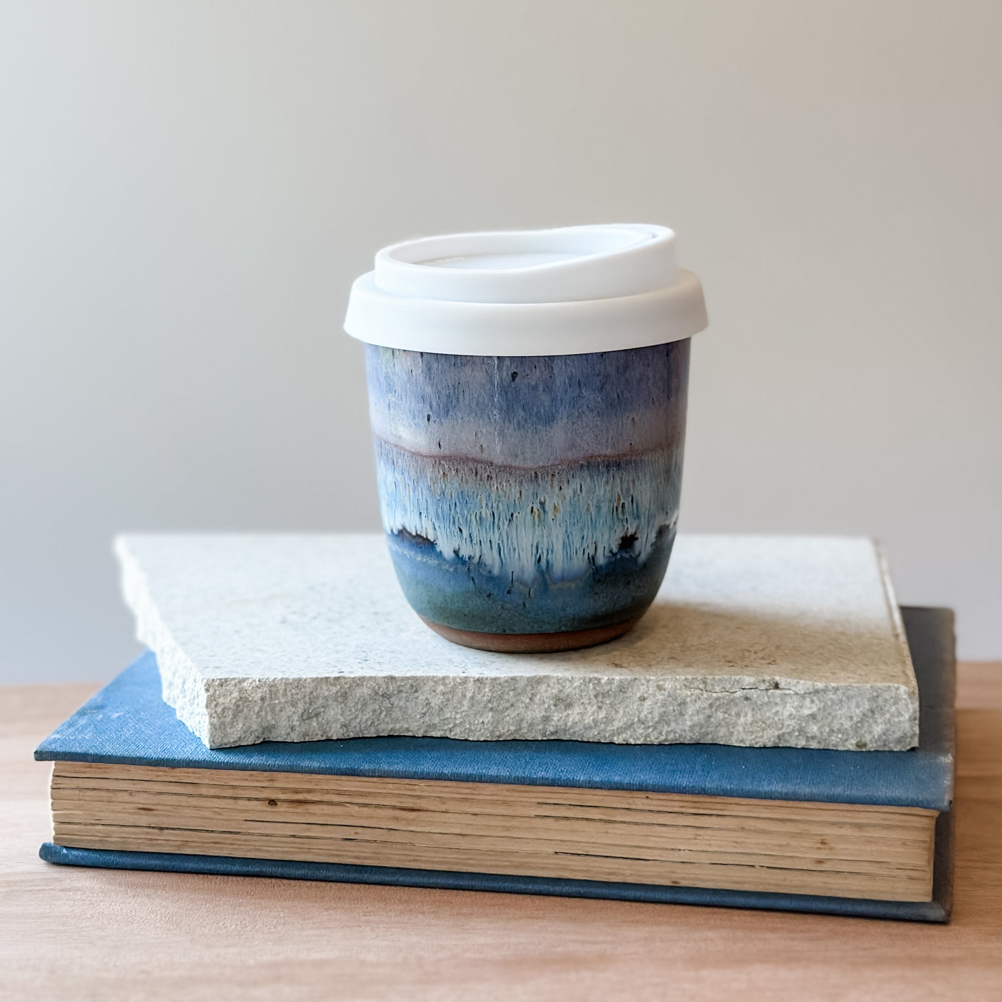 Purple, white, and blue ceramic keep-cup with a white lid on top of books against a plain background