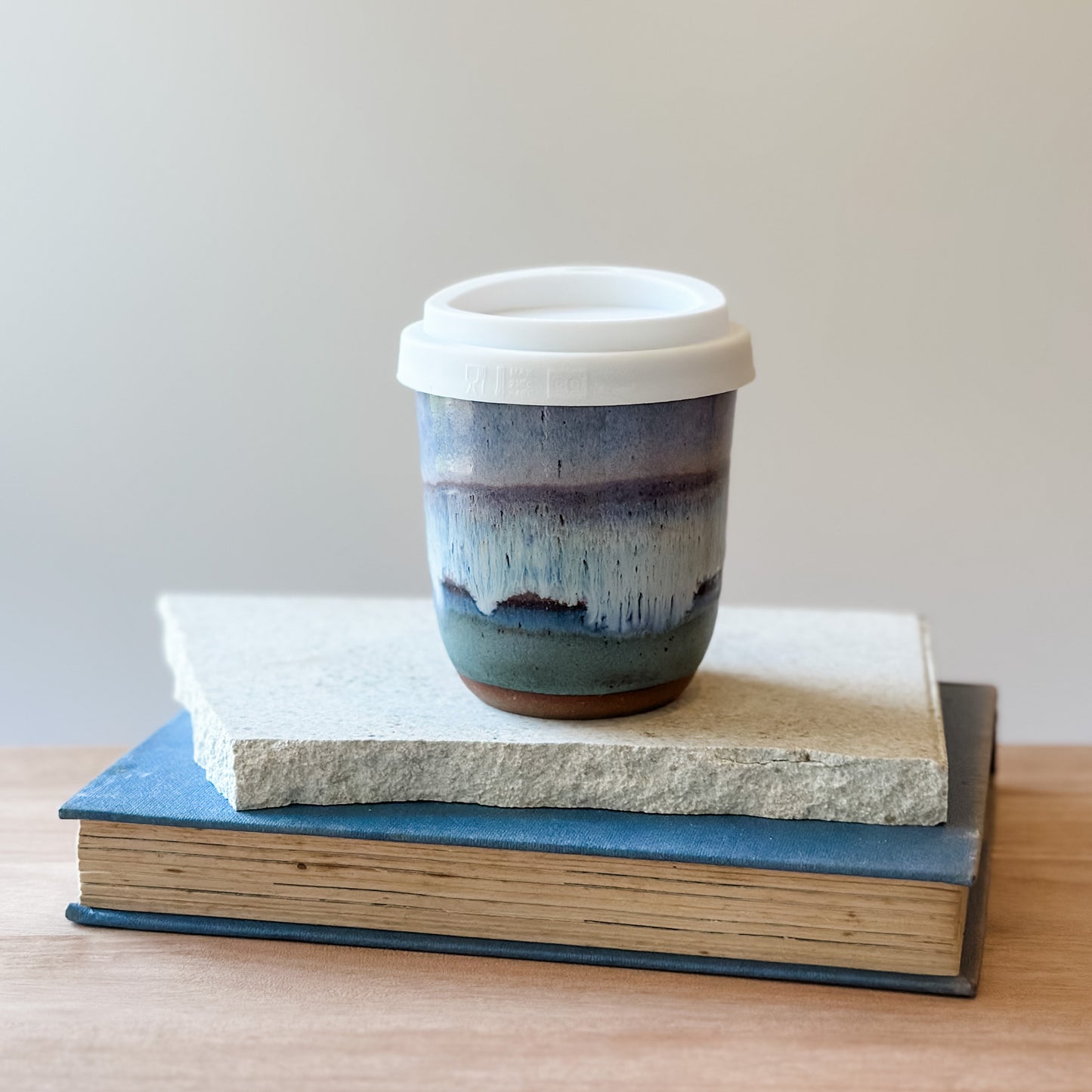 Ceramic cup with a white lid on top of a tile and a book on a wooden surface.