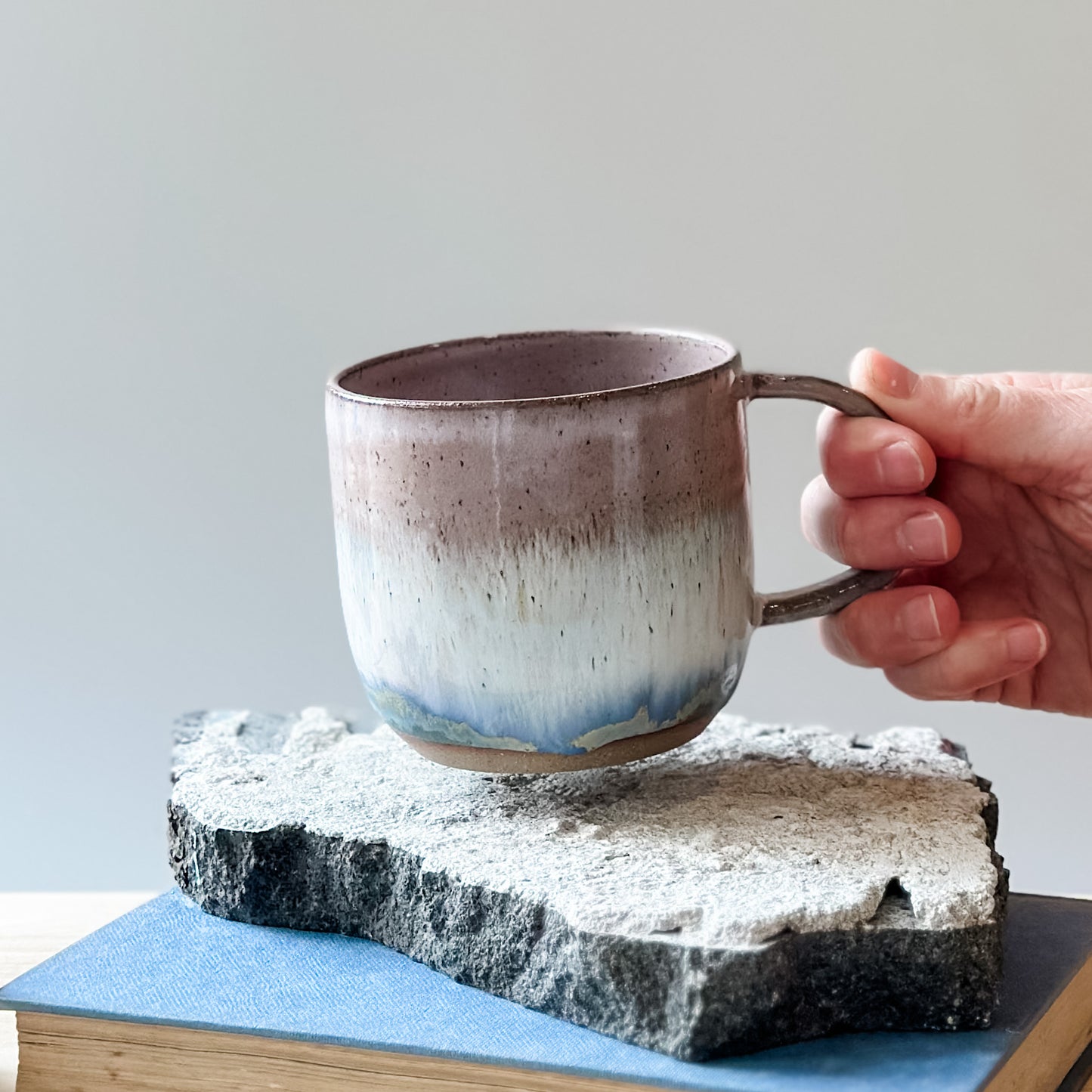 A handmade ceramic mug in blush pink, white and blue glaze. The handmade mug is being held by the handle above a tile.