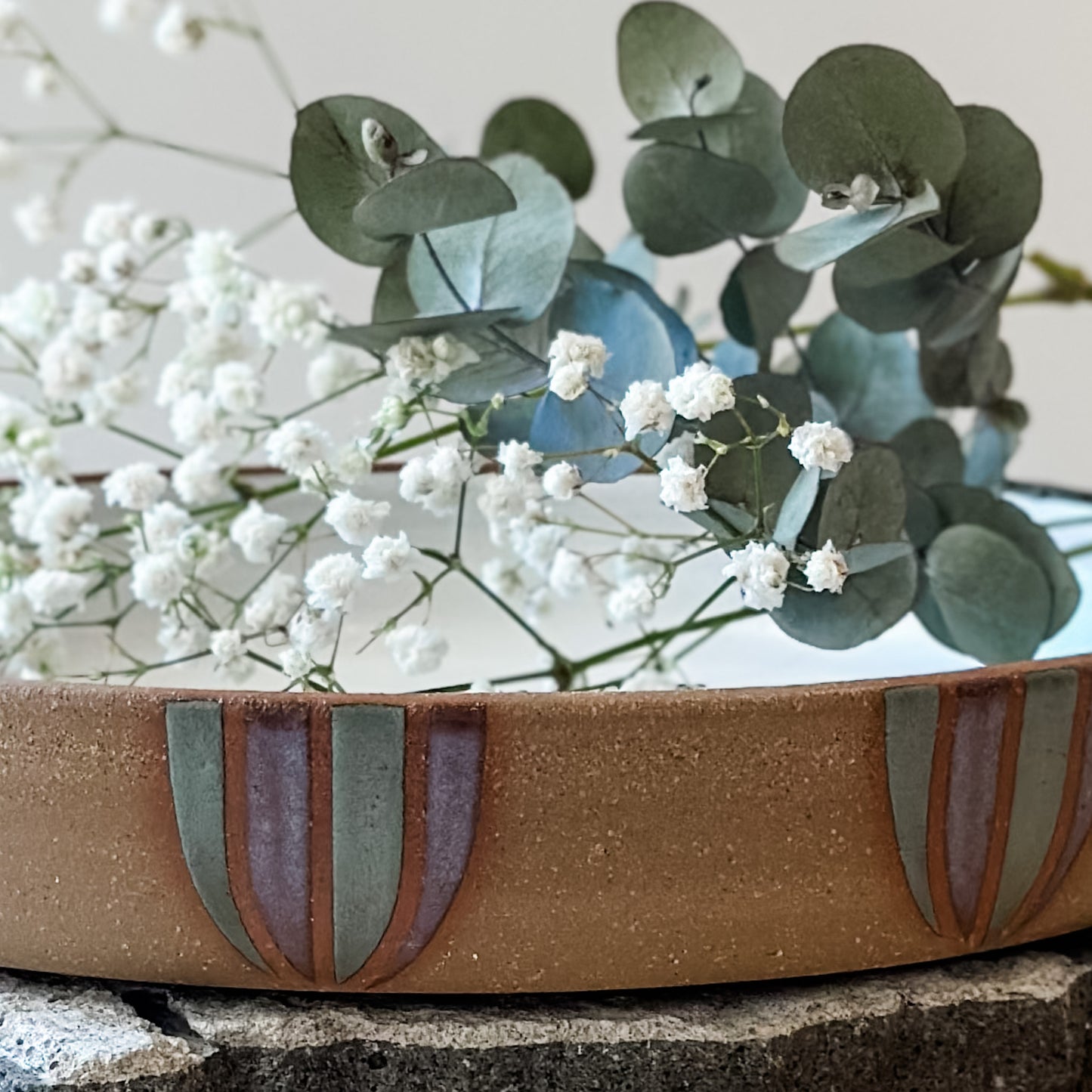 A close-up view of a Coastal Handmade Ceramic Platter on a tile with flowers. The platter has a white interior and a purple and blue-grey design on the exterior.