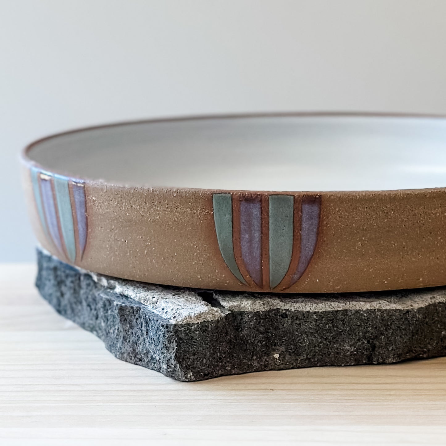 A close-up view of a Coastal Handmade Ceramic Platter on a tile and a wooden table. The platter has a white interior and a purple and blue-grey design on the exterior.