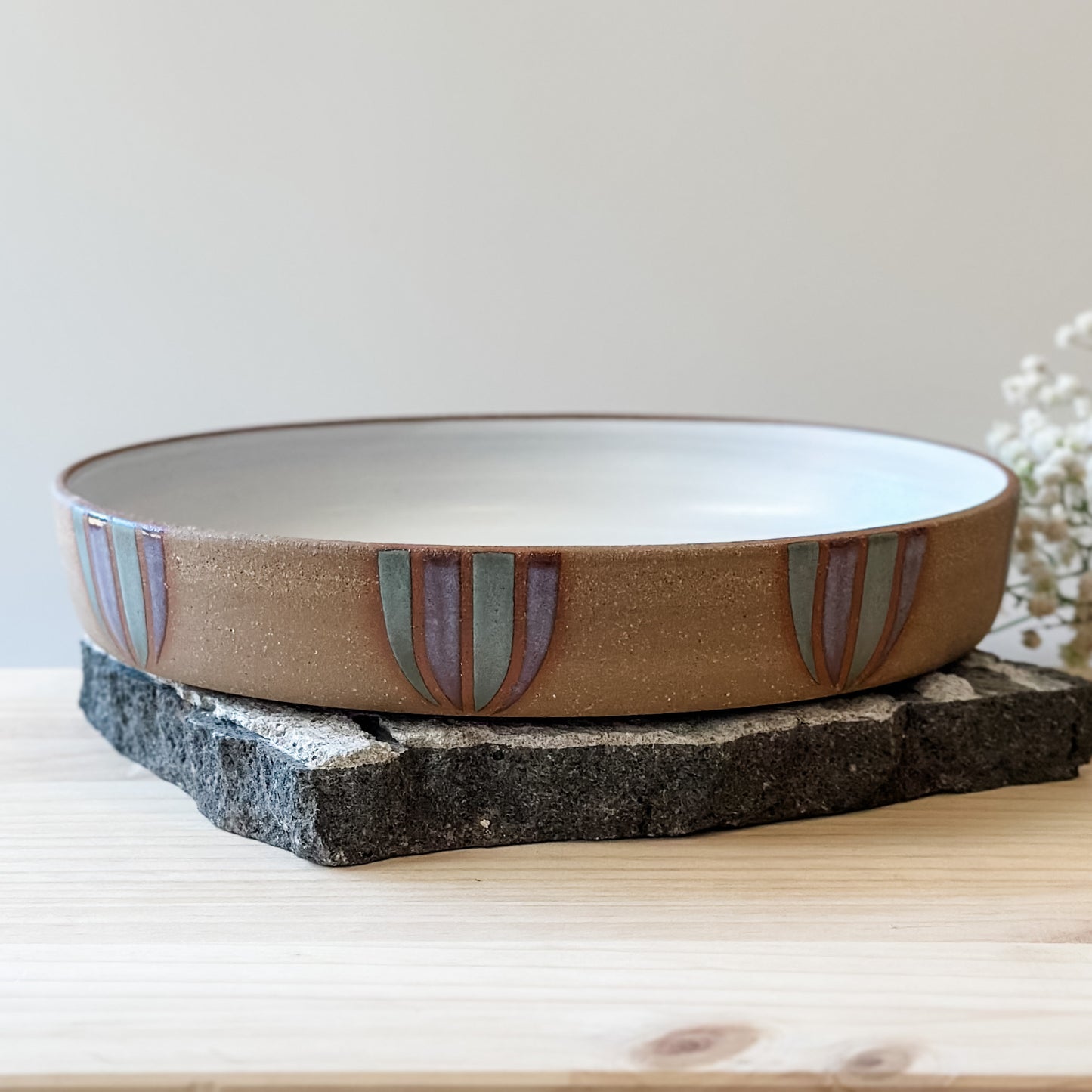 A front view of a Coastal Handmade Ceramic Platter on a tile and a wooden table. The platter has a white interior and a purple and blue-grey design on the exterior.