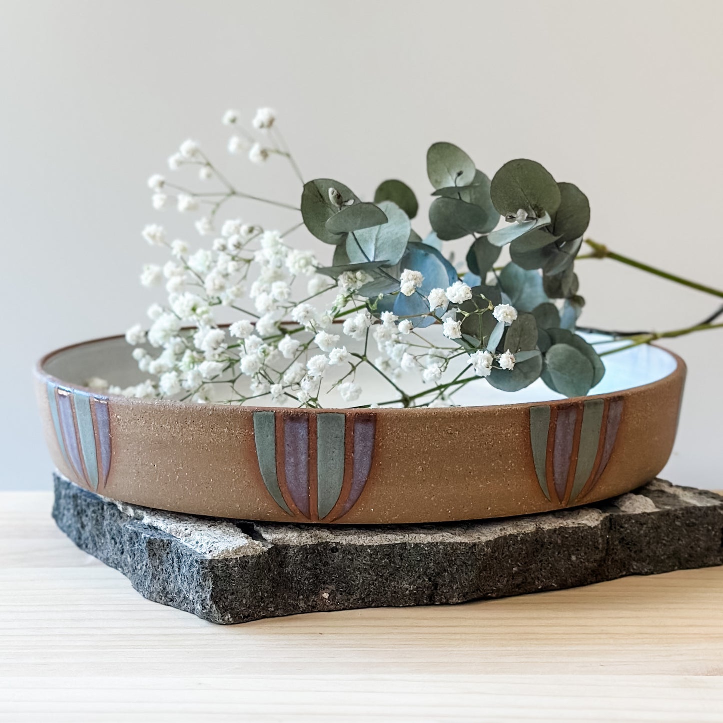 A front view of a Coastal Handmade Ceramic Platter with flowers on a tile and on a wooden table. The platter has a white interior and a purple and blue-grey design on the exterior.