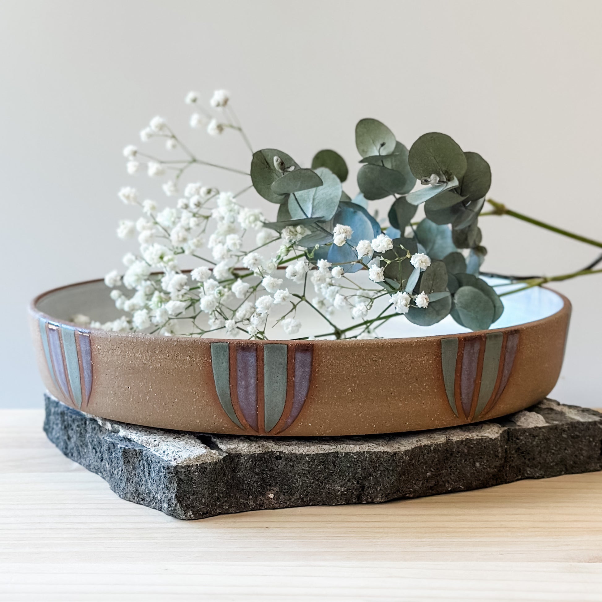 A front view of a Coastal Handmade Ceramic Platter with flowers on a tile and on a wooden table. The platter has a white interior and a purple and blue-grey design on the exterior.