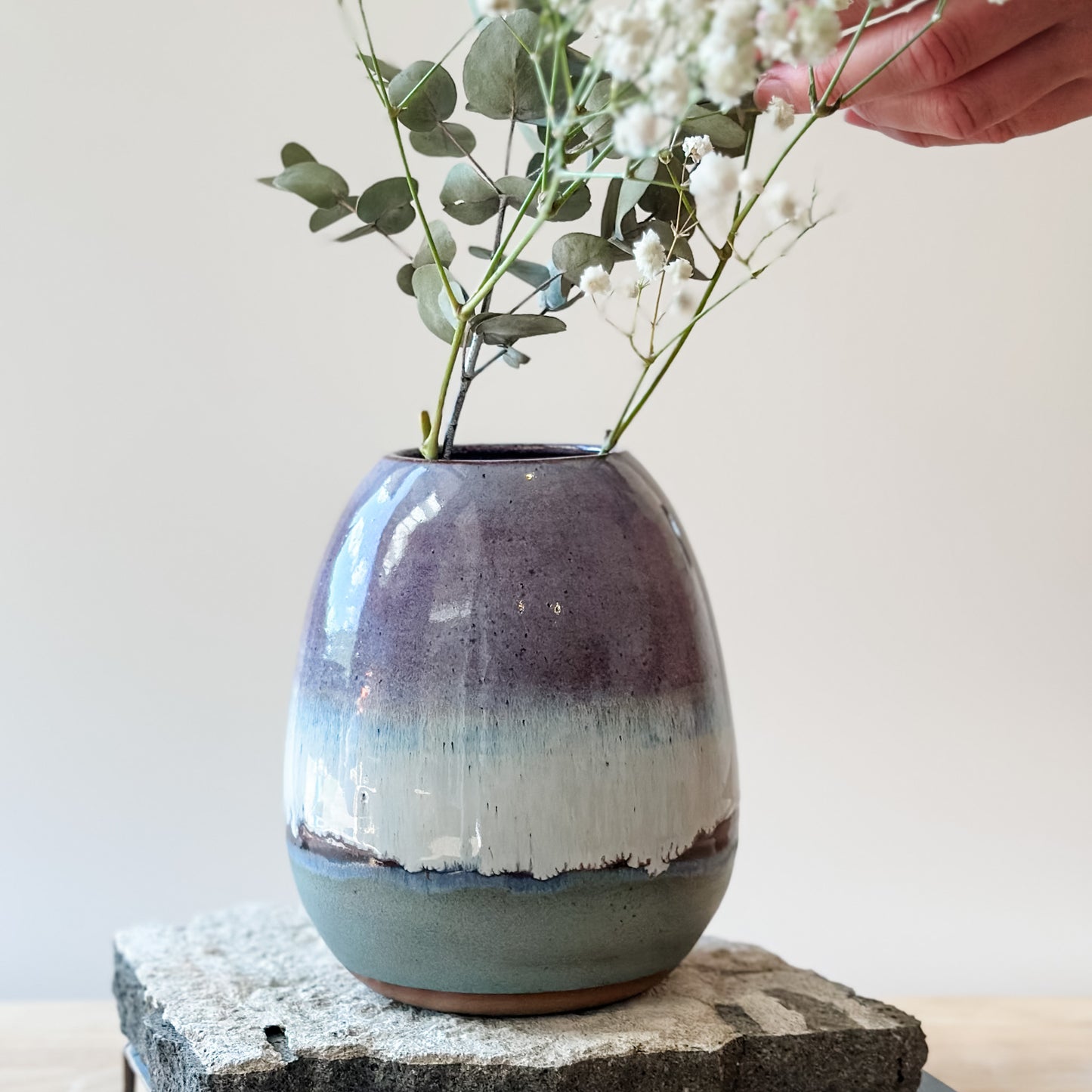 The Coastal Handmade Ceramic Round Vase sits on a tile and wooden table. The vase has a purple, white and blue glaze. A hand is arranging gum leaves and baby's breath flowers.