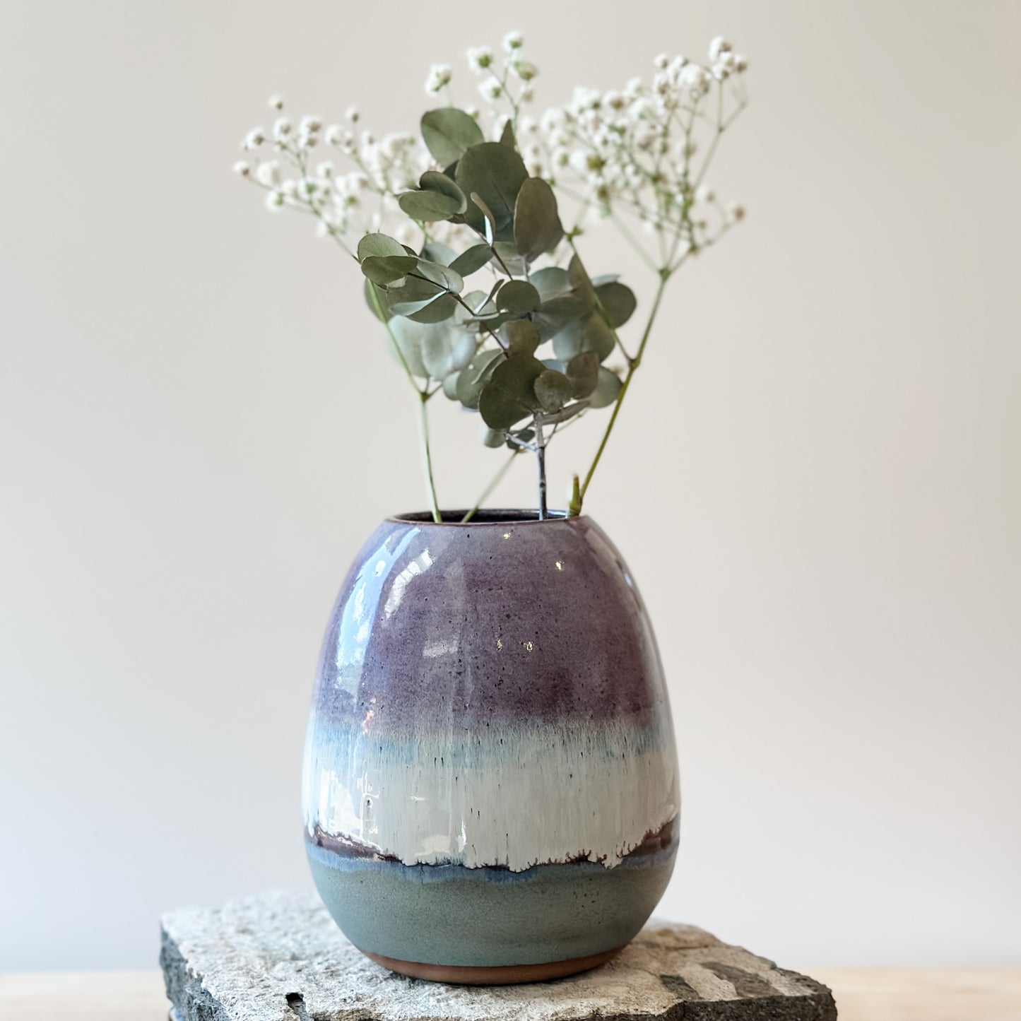 The Coastal Handmade Ceramic Round Vase sits on a tile and wooden table. The vase has a purple, white and blue glaze. The ceramic vase holds gum leaves and baby's breath flowers.