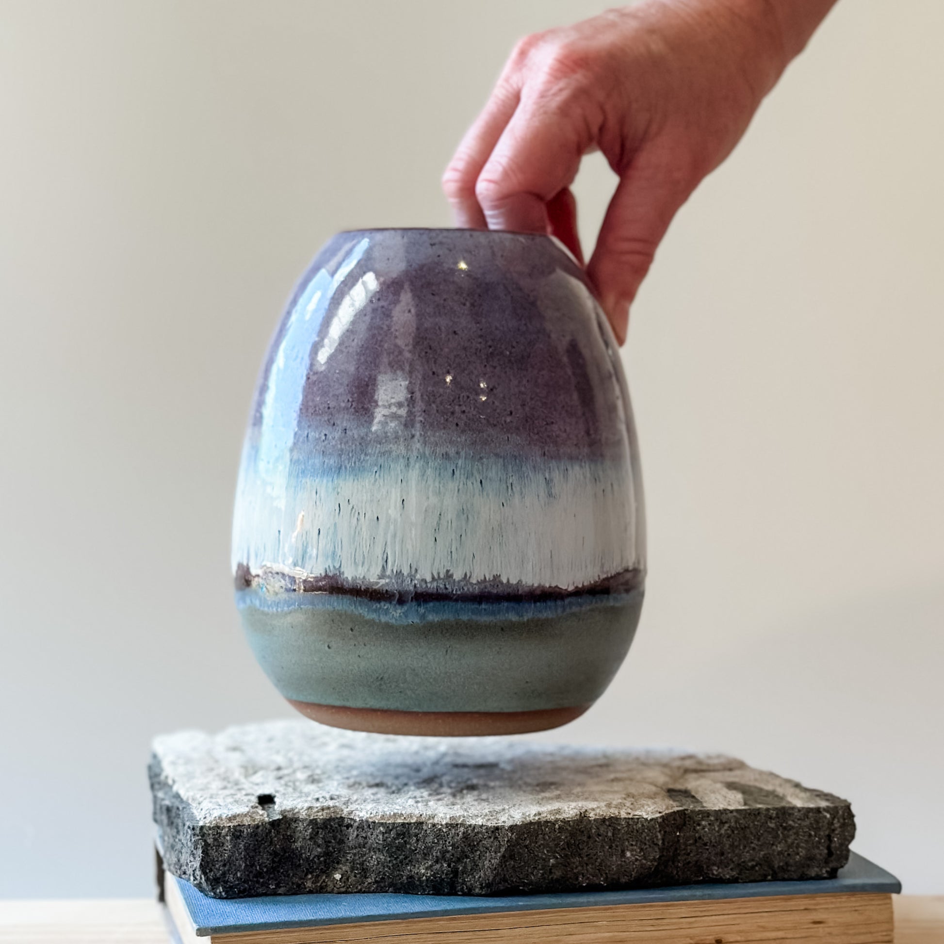 The Coastal Handmade Ceramic Round Vase is being on a tile and wooden table. The vase has a purple, white and blue glaze.